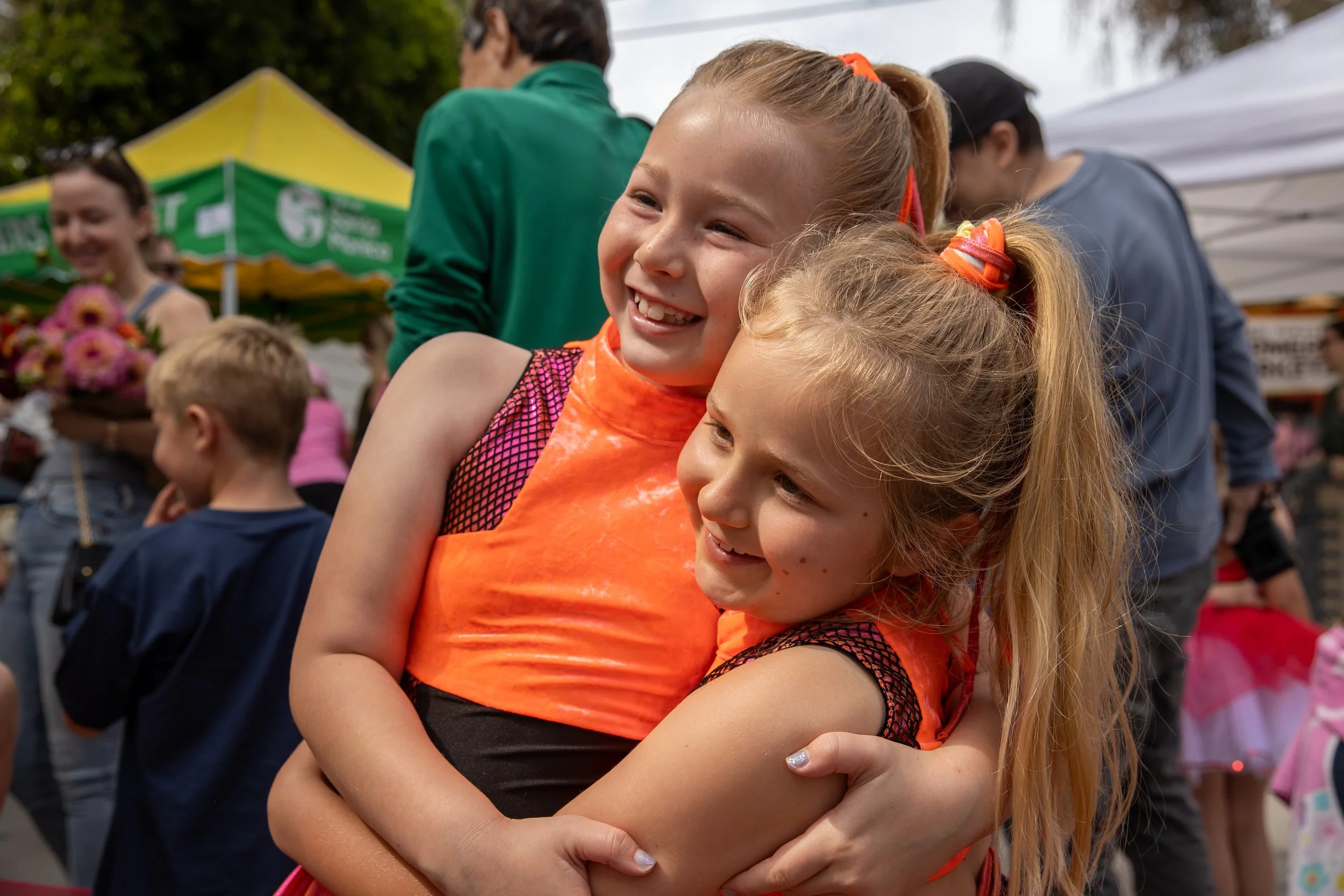 Two young girls smiling and hugging at an outdoor event, surrounded by people and colorful tents.