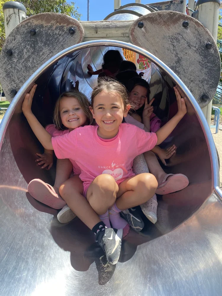 Four children smiling and sitting inside a metal playground tube slide at a park, with a clear sky and green trees in the background.