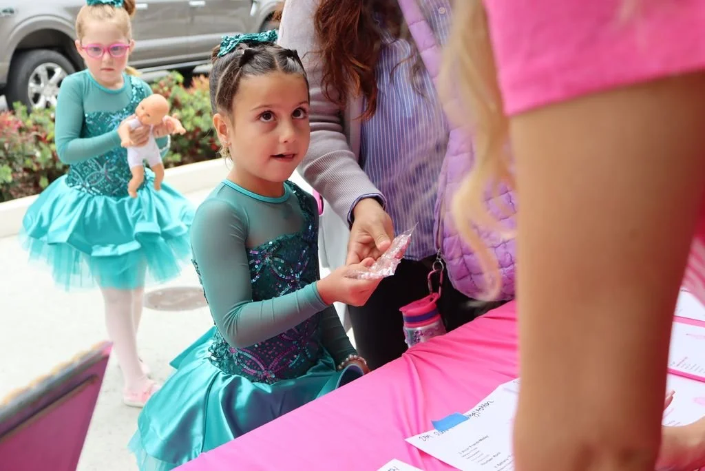 A young girl in a blue ballet costume holding a small plastic-wrapped object, talking to another person at a pink table during an outdoor event. Another girl in a similar costume, holding a doll, is in the background.