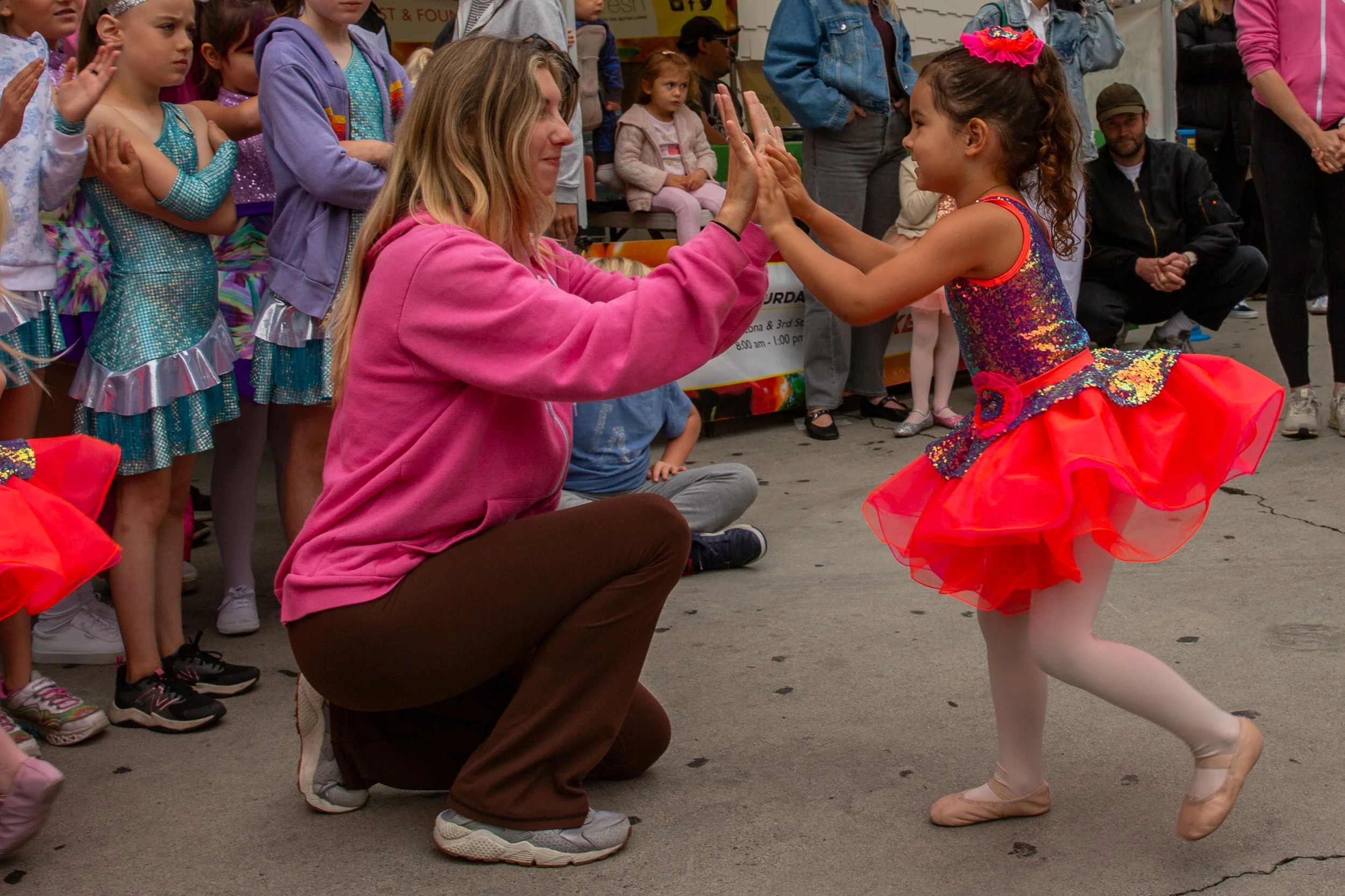 A young girl in a colorful, sparkly dress and tutu is dancing and high-fiving a woman in a pink sweatshirt, surrounded by other children and onlookers at an outdoor event.