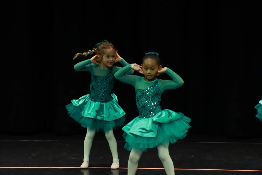 Two young girls in teal dance costumes perform on stage, with black background and stage floor.