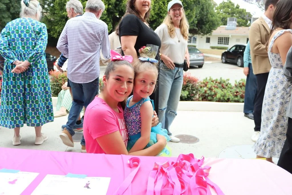 Two young girls sitting at a pink table with ribbons, smiling, as people gather outside in a park-like setting for an event or celebration.