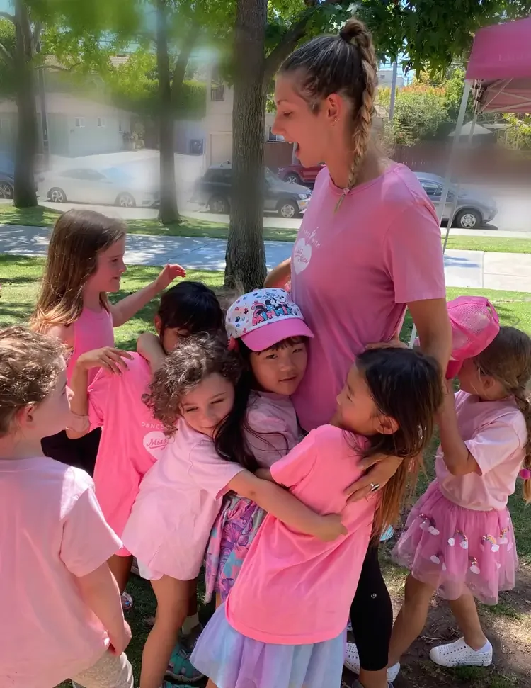 Young woman and children, all wearing pink, sharing a group hug outdoors under a tree