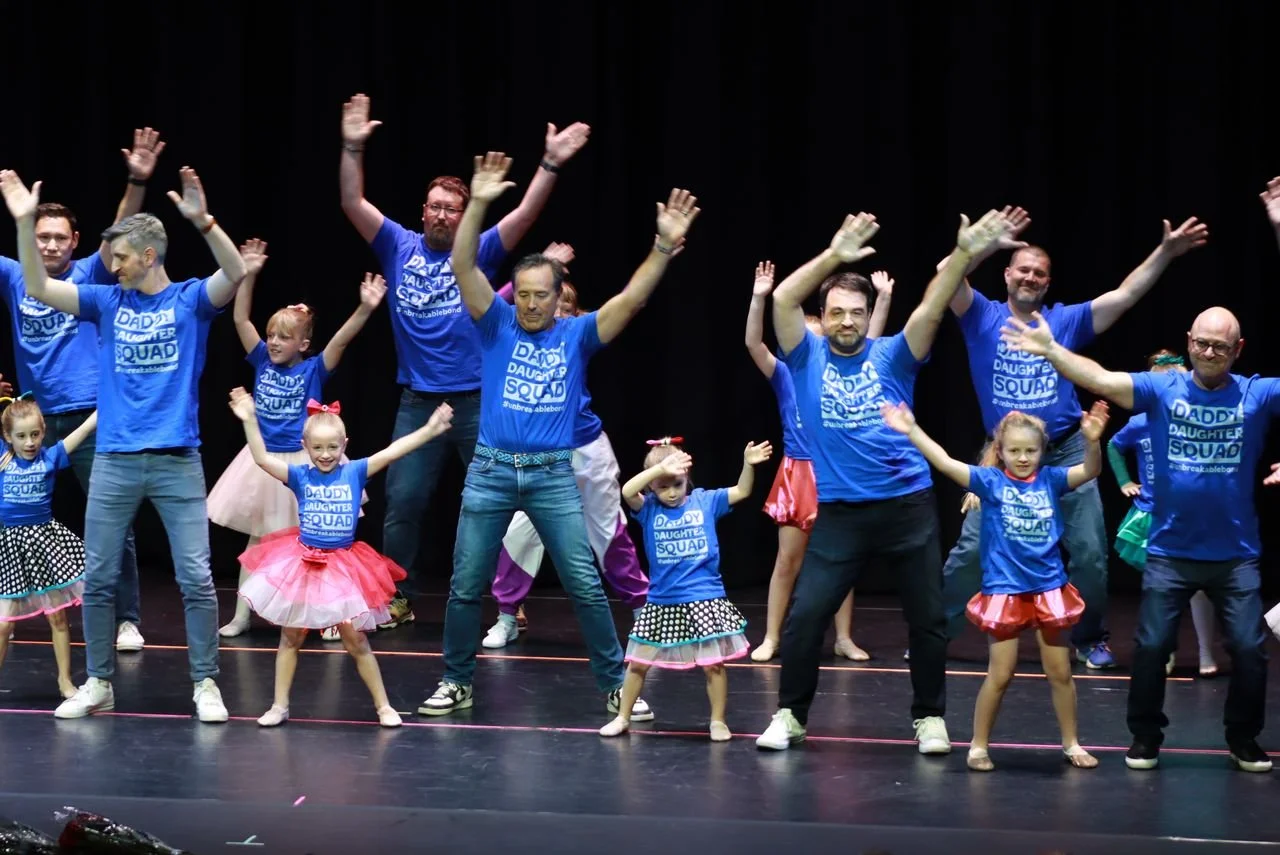 Group of adults and children dancing on stage, all wearing matching blue T-shirts that say "Daddy Daughter Squad," with the adults raising their hands and the children happily participating.
