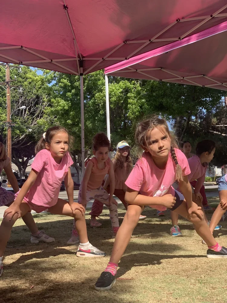 Group of young girls in pink shirts doing a workout or dance under a pink canopy in a park.
