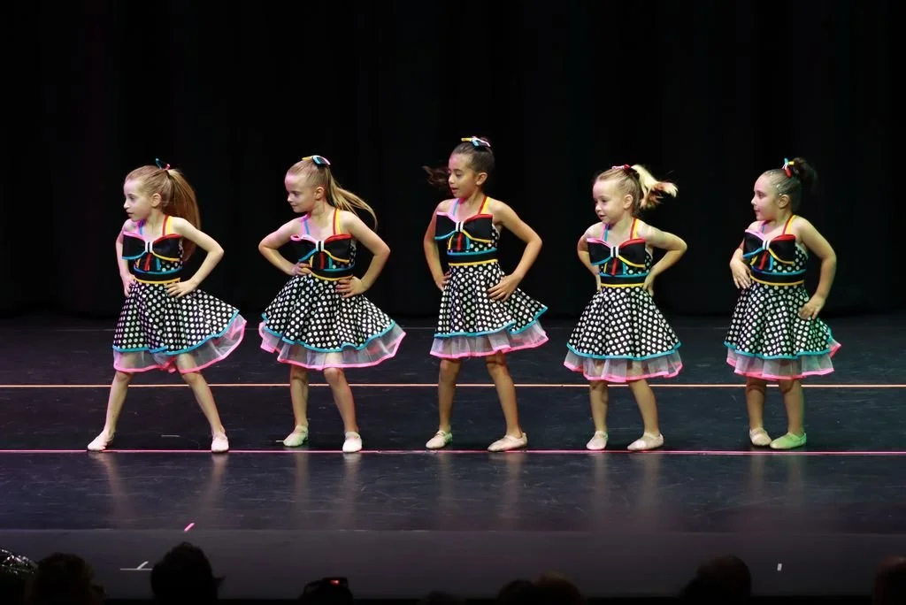 Five young girls in colorful polka dot dresses performing a dance on stage.