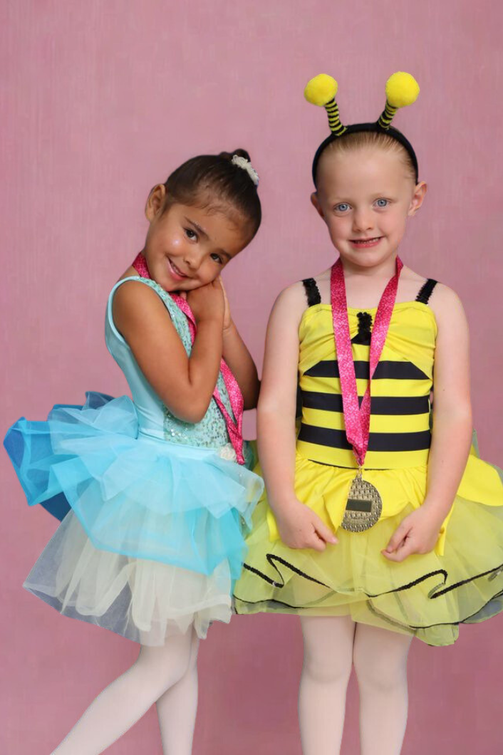 Two young girls dressed in colorful costumes standing against a pink background. One girl is wearing a light blue tutu dress and is posing with a shy smile, while the other girl is dressed as a bee with a yellow and black striped dress, bee antenna h
