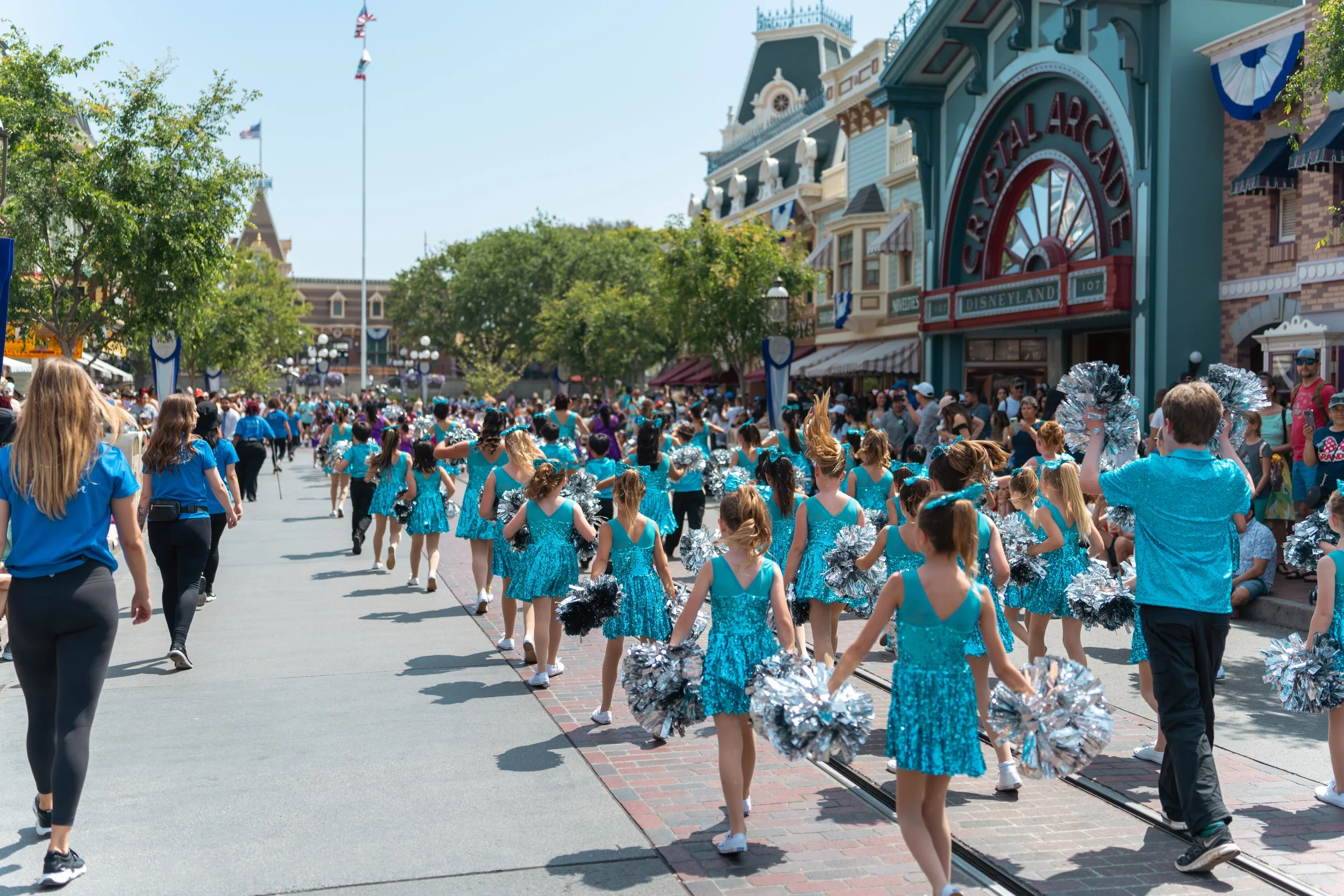 Group of young cheerleaders in blue outfits with pom-poms performing a parade at Disneyland with the entrance in the background.