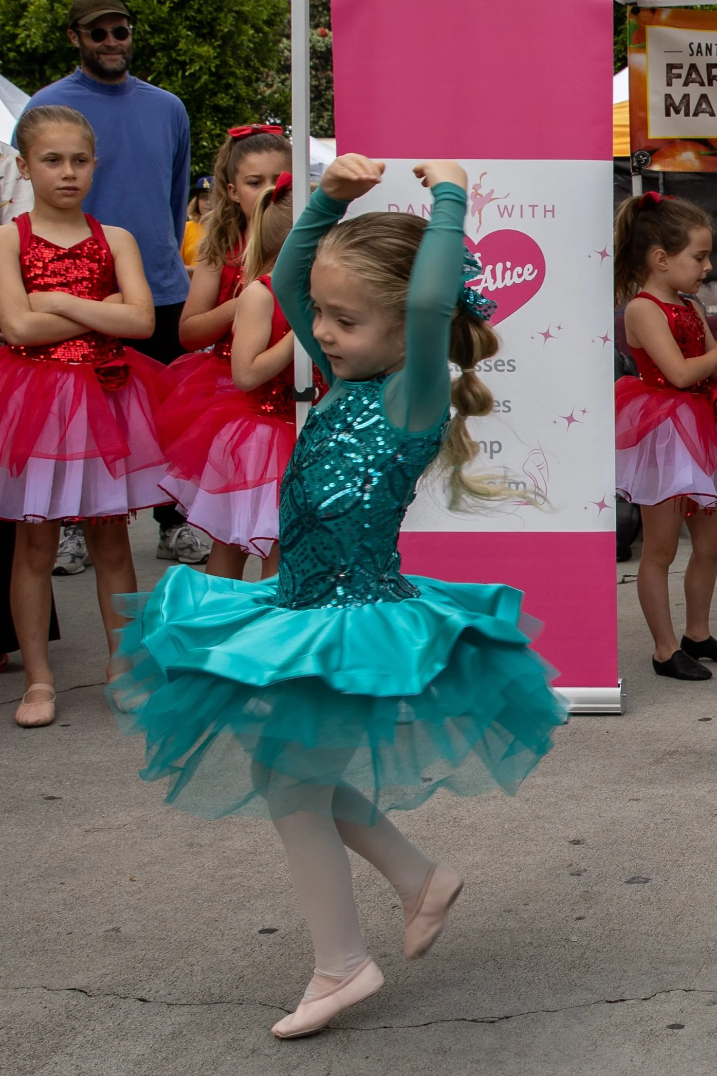 Young girl wearing a turquoise ballet costume performing a dance move at an outdoor event, with other girls in red costumes and a man in a blue shirt observing in the background.