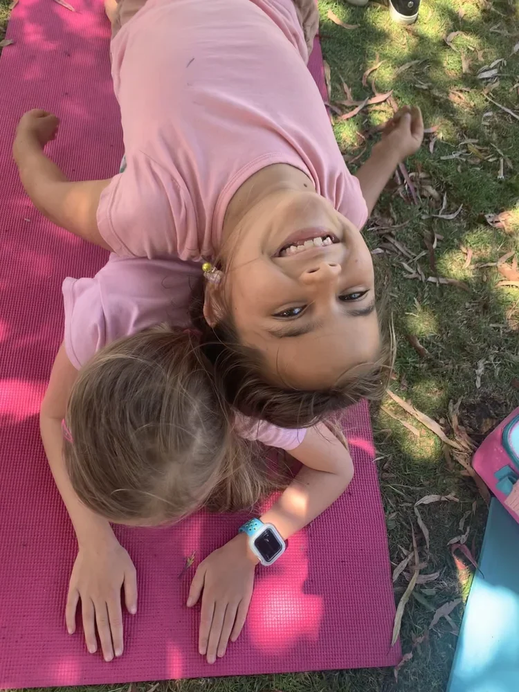 Two young girls lying on a pink yoga mat outdoors, one smiling at the camera and the other with head resting on the other's back, surrounded by grass and fallen leaves.