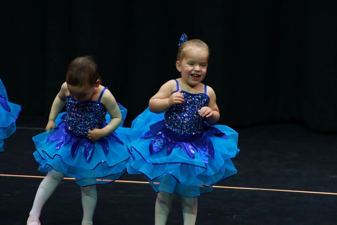 Two young girls in blue sparkly tutu costumes performing on stage, one smiling and the other looking down, against a black background.