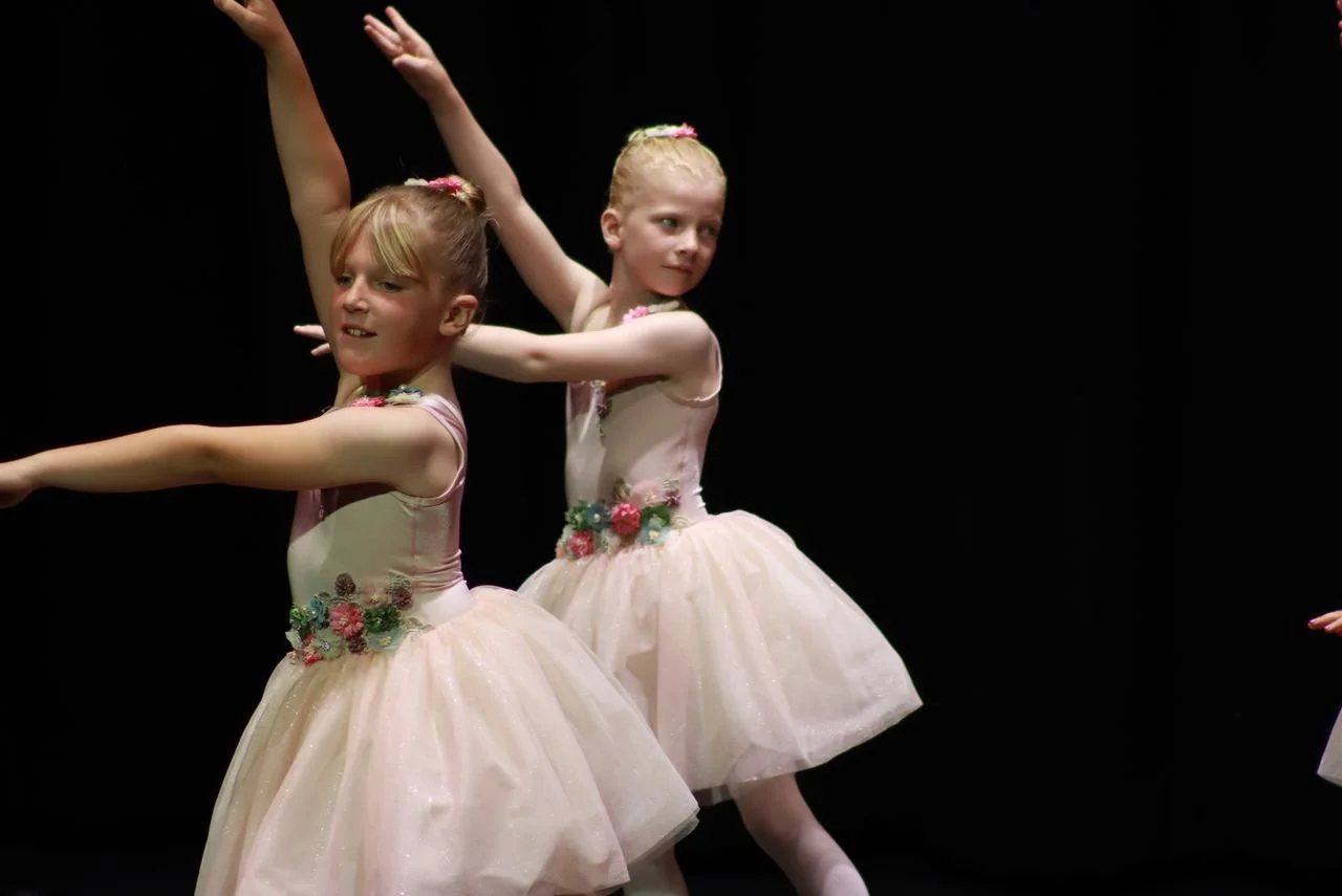 Two young girls in ballet costumes performing a dance on stage against a black background.