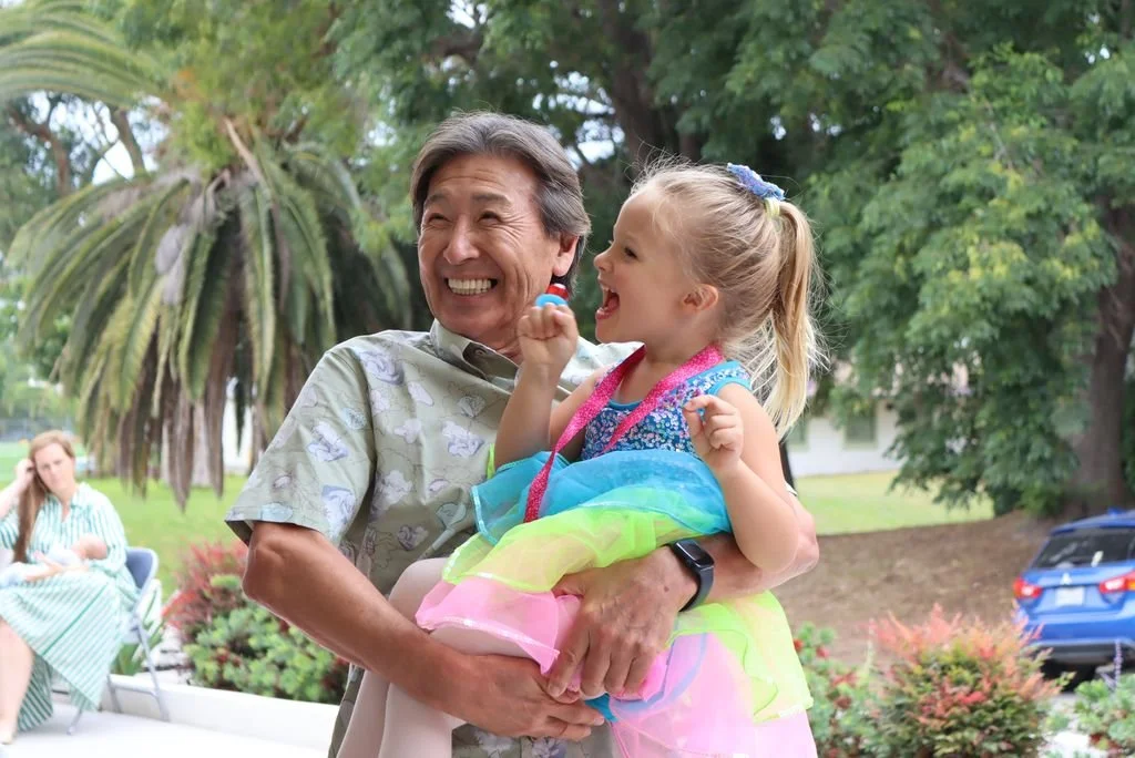 A joyful elderly man holding a young girl in a colorful dress outdoors, with a woman sitting on a chair in the background and trees surrounding them.
