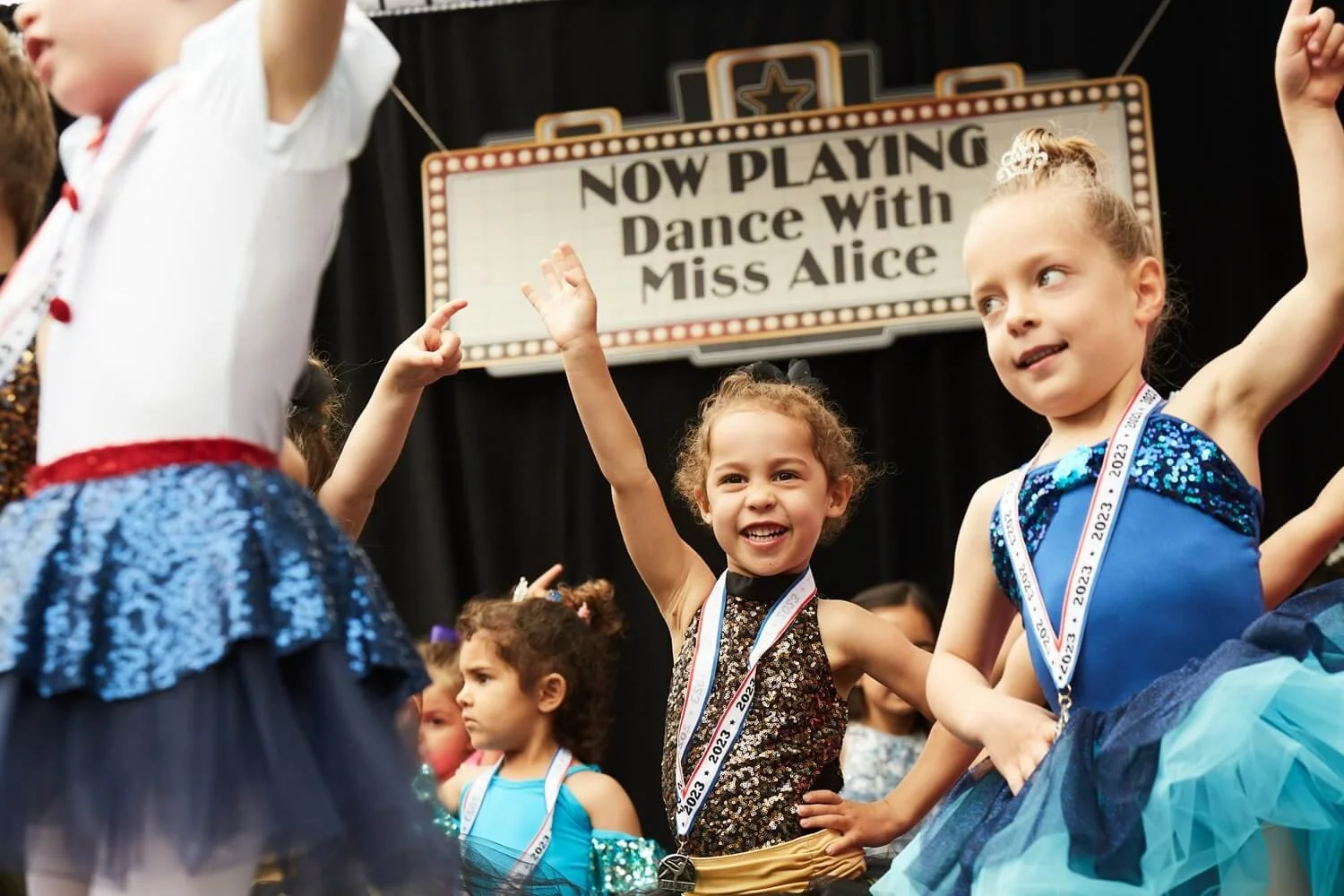 Young children wearing dance costumes and medals participating in a dance competition with a sign that reads 'Now Playing: Dance With Miss Alice' in the background.