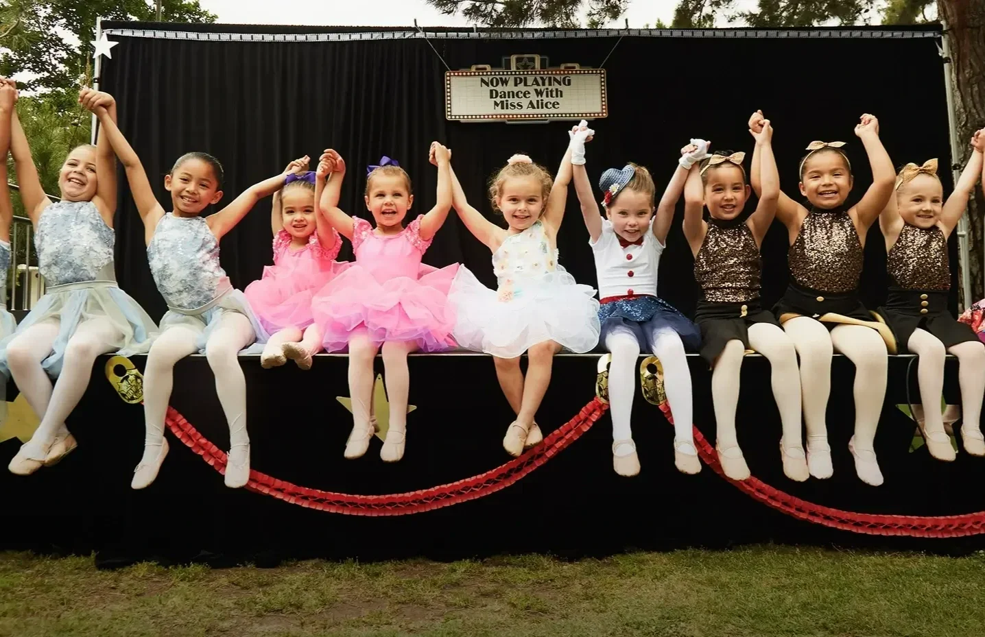 Students of Dance with Miss Alice dressed in colorful dance costumes sitting on a stage, holding hands, with a sign above that reads "Now Playing Dance With Miss Alice."