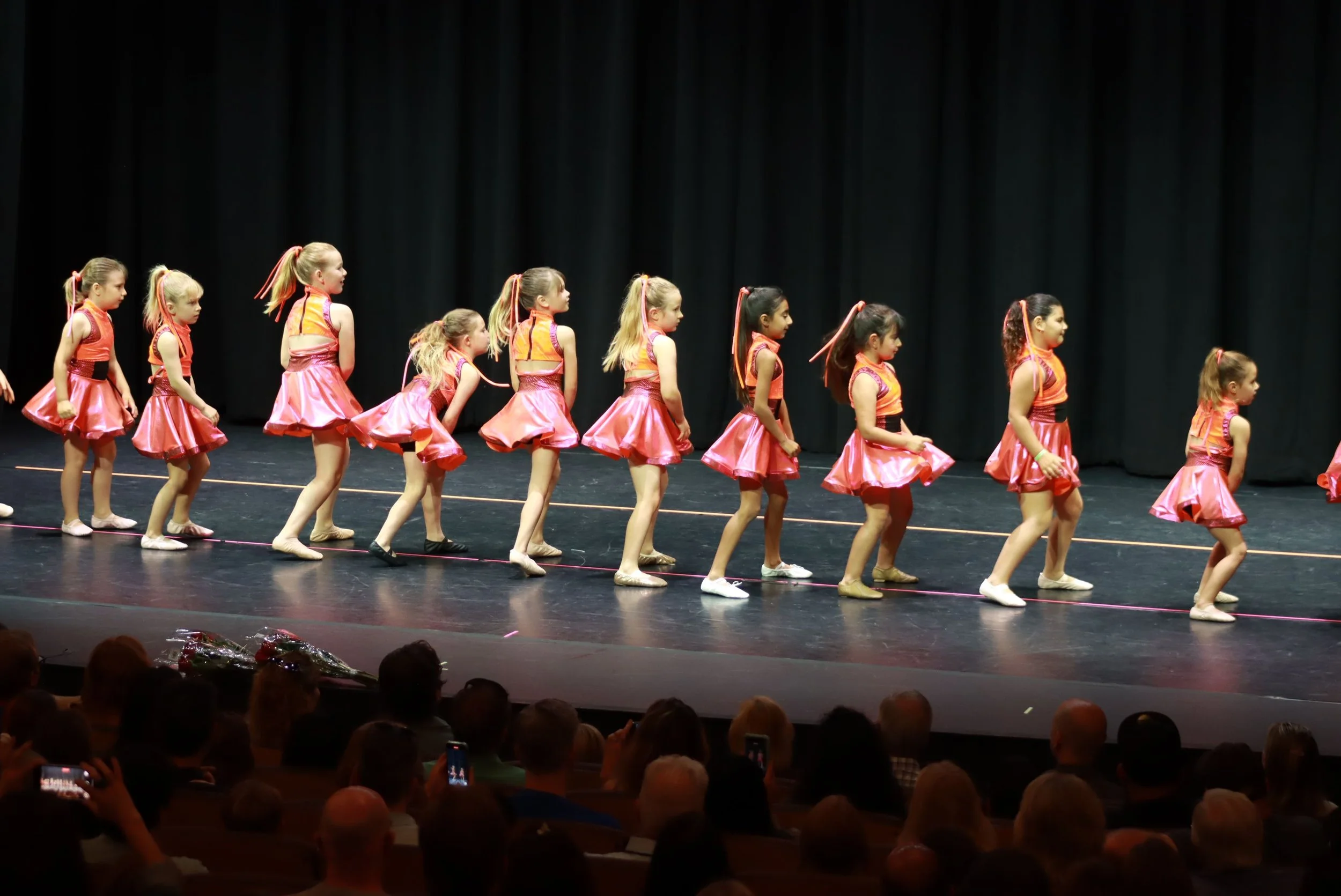 A line of young girls in pink costumes performing on stage during a dance recital, with an audience watching.