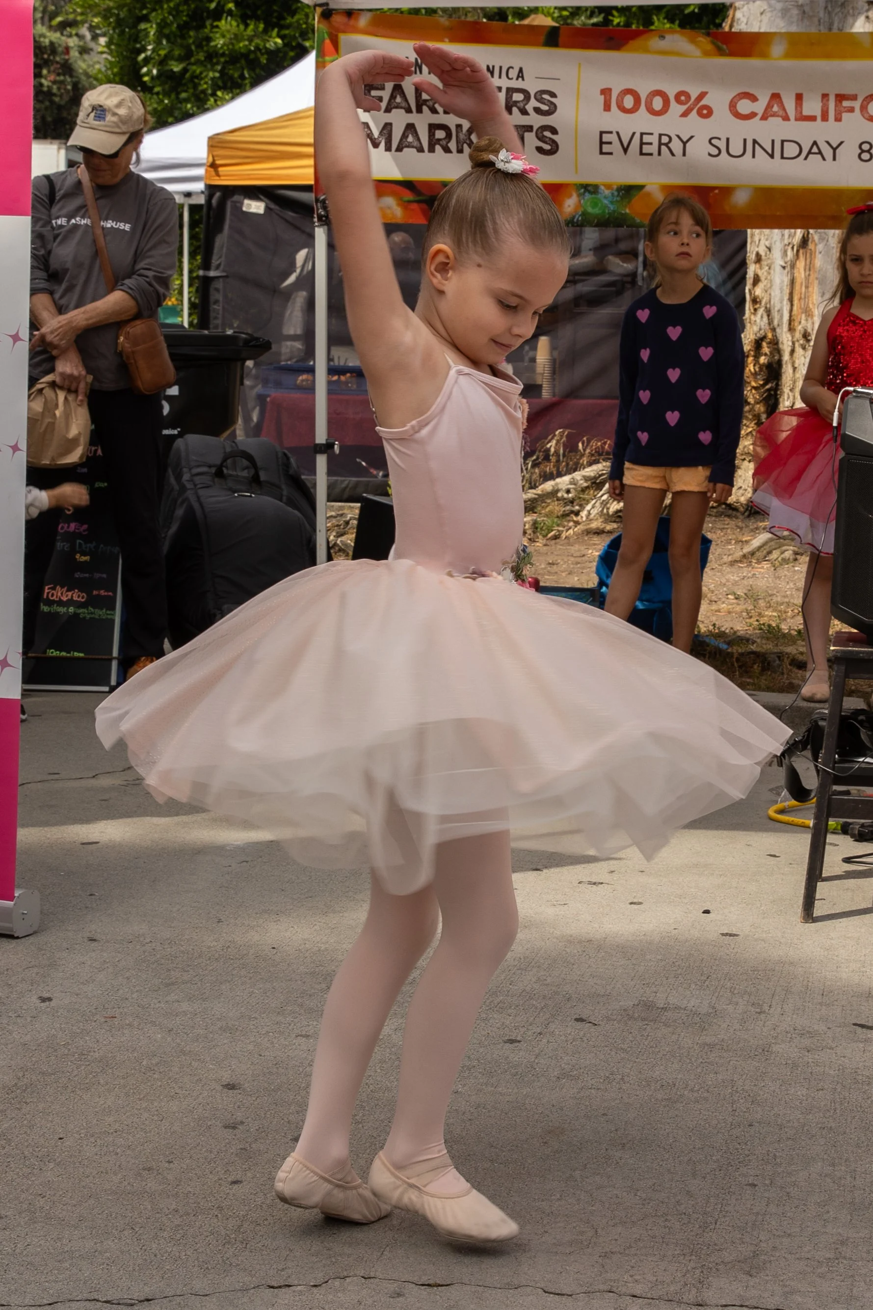 A young girl dressed in a ballerina tutu and tights is performing a dance at an outdoor event. She has her arms raised above her head and appears to be in motion.