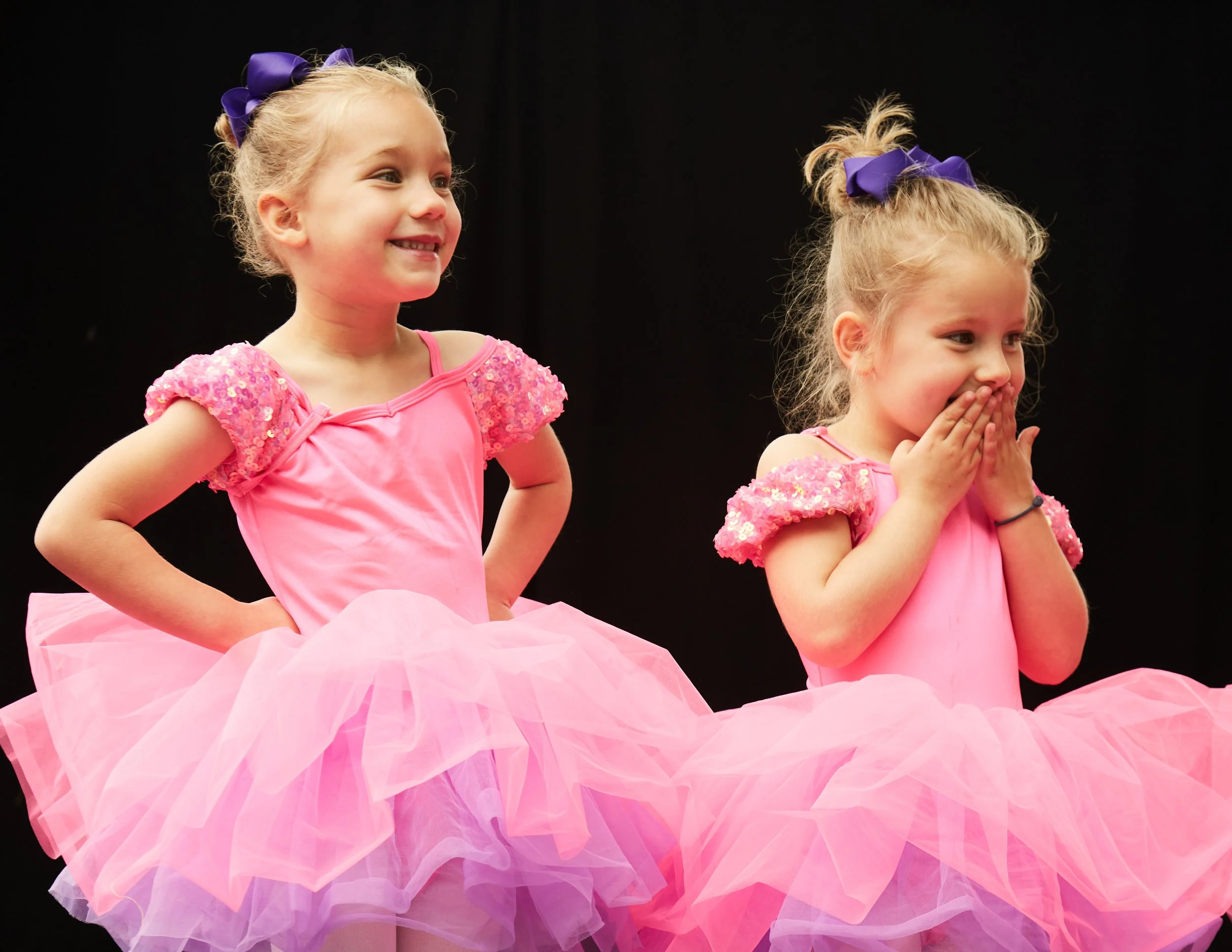 Two young girls dressed in pink ballet tutus and matching tops with sequin sleeves, smiling and covering her mouth with her hands, standing on a dark background.