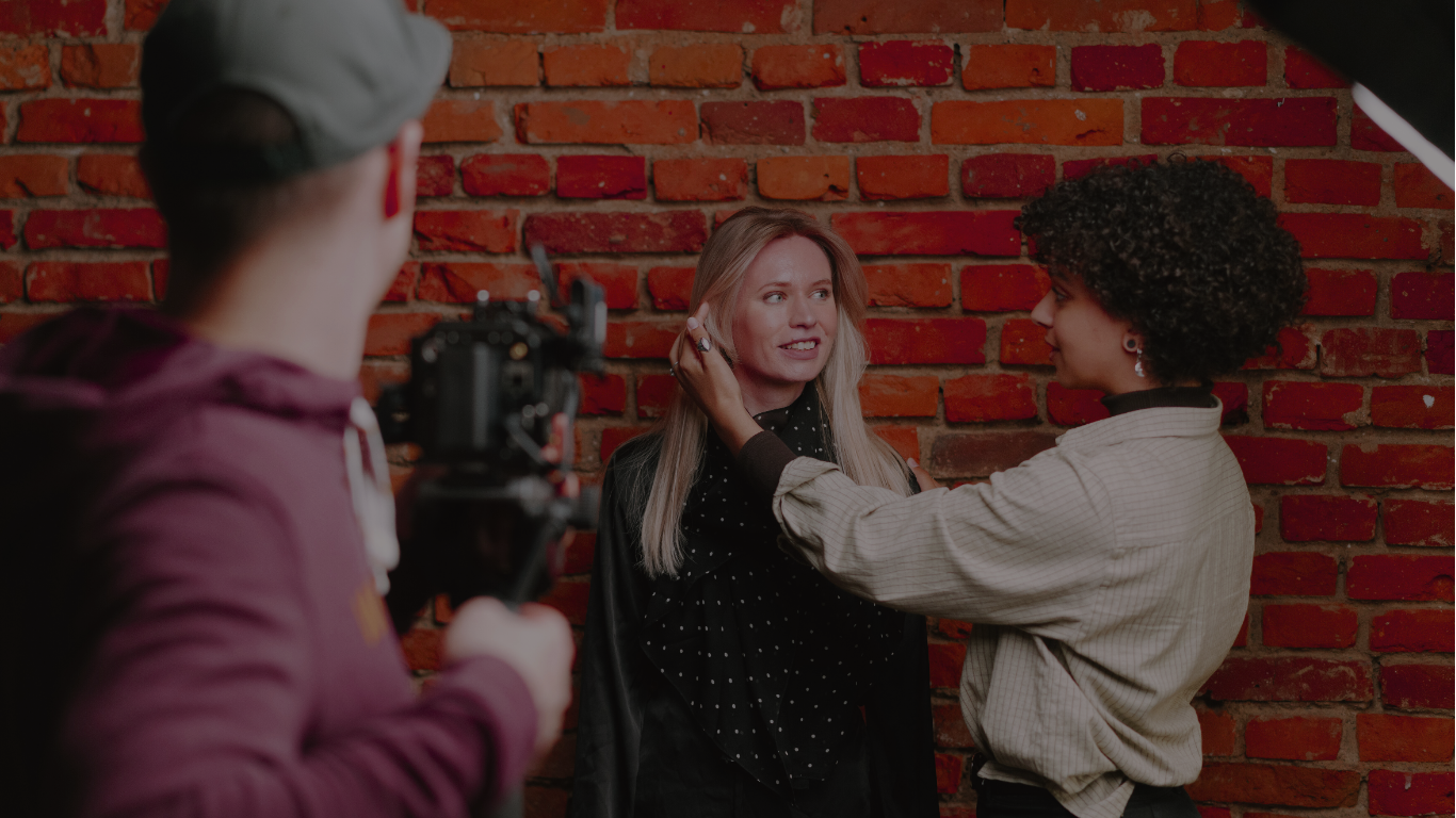 A woman with long blonde hair has makeup applied by a woman with short curly hair against a red brick wall, while a person with a camera records the scene.