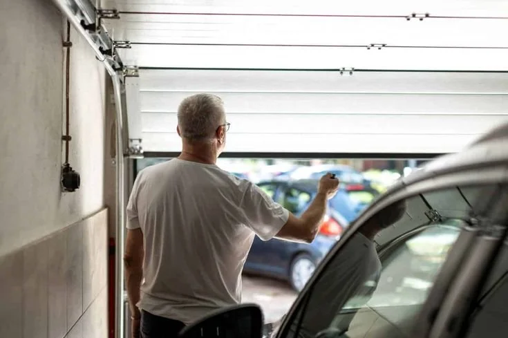 A man with gray hair wearing a white t-shirt is seen from behind, standing inside a garage and looking outside through an open garage door, with parked cars visible outside.
