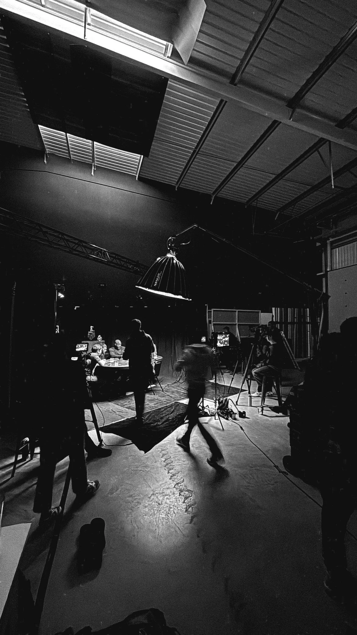 Black and white image of a film or TV show production set with crew members, cameras, and lighting equipment, and a tabletop scene with actors or performers.