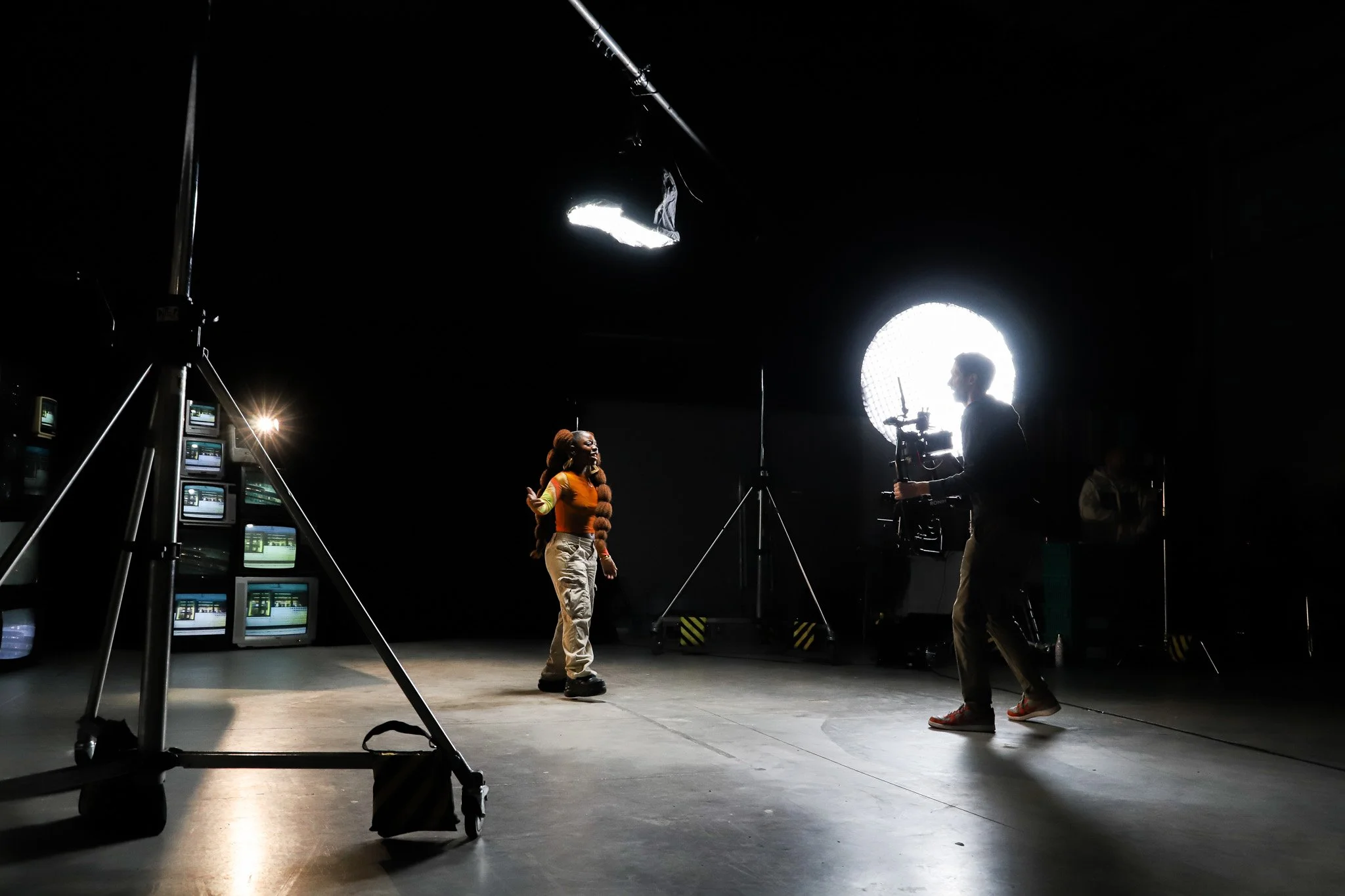 Film set with two people, one woman talking, surrounded by professional lighting and monitors, inside a dark studio.