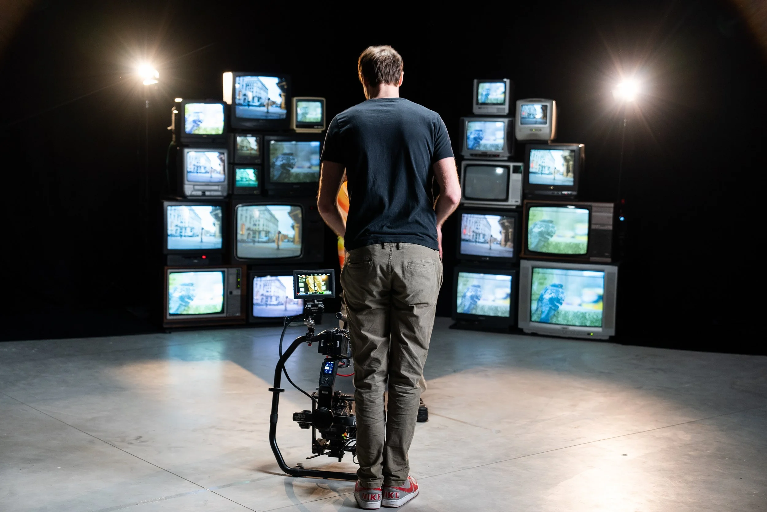 A person wearing grey pants and a black t-shirt standing in a room with many old television screens displaying various images, with bright stage lights in the background.