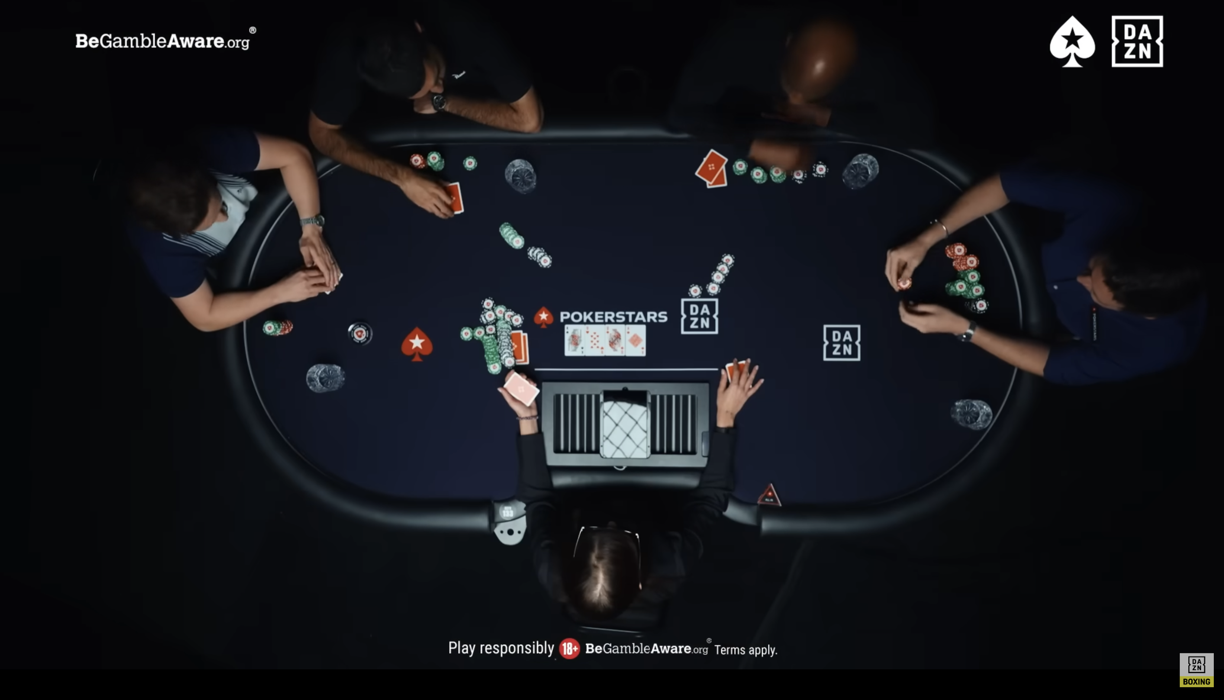 An overhead view of a poker game at a black table with multiple players. Players are handling poker chips and cards. The scene includes branding for BeGambleAware.org and DAZN, with a logo for PokerStars. The dealer is centrally positioned with a laptop and hands dealing cards. The atmosphere suggests a professional or televised poker game.
