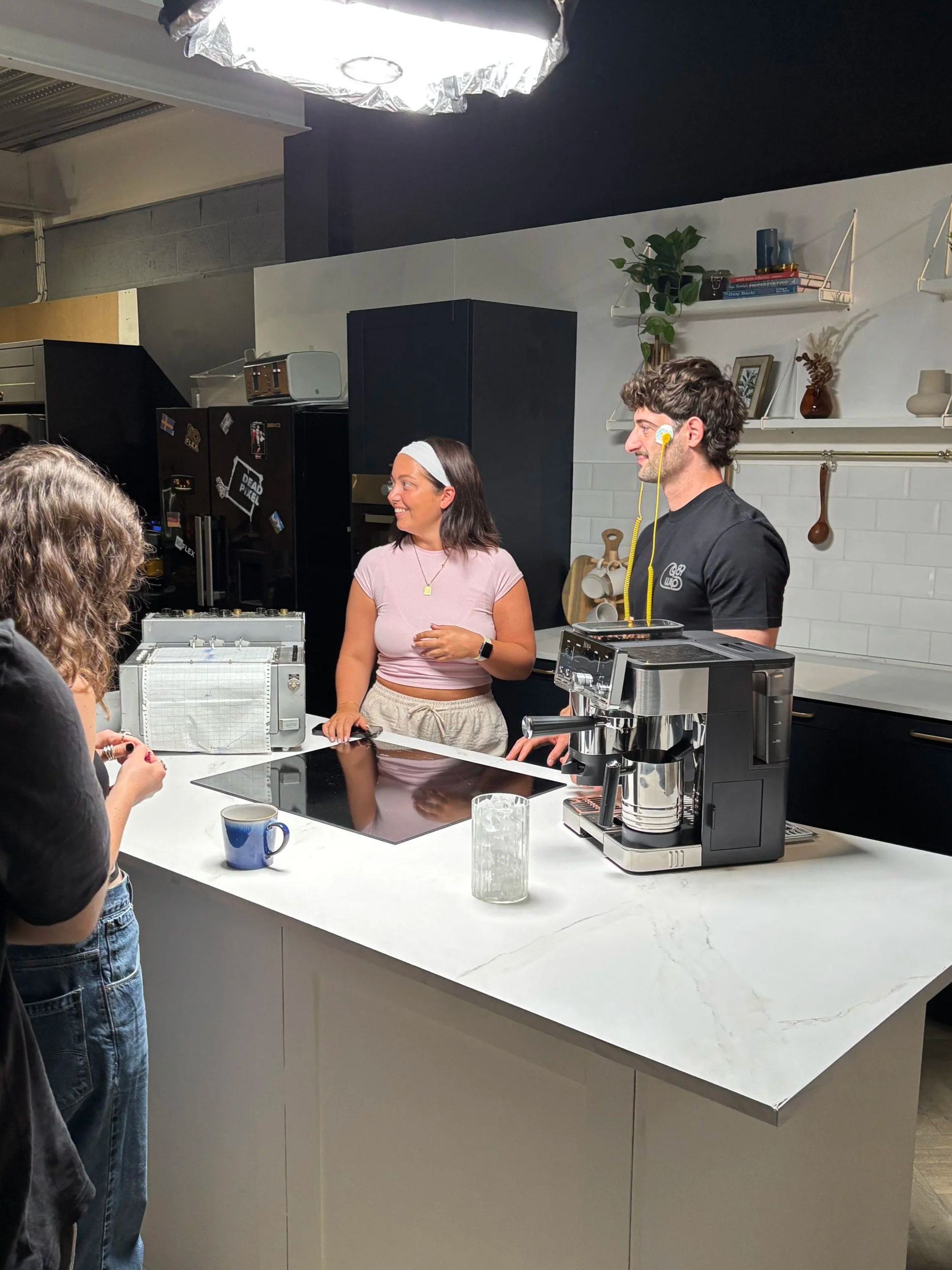 Two people are standing in a modern kitchen, a woman with dark hair and a woman with a yellow ear and black shirt, talking to two women whose faces are not visible. The kitchen has a white marble countertop, a coffee machine, and shelves with decorative items.