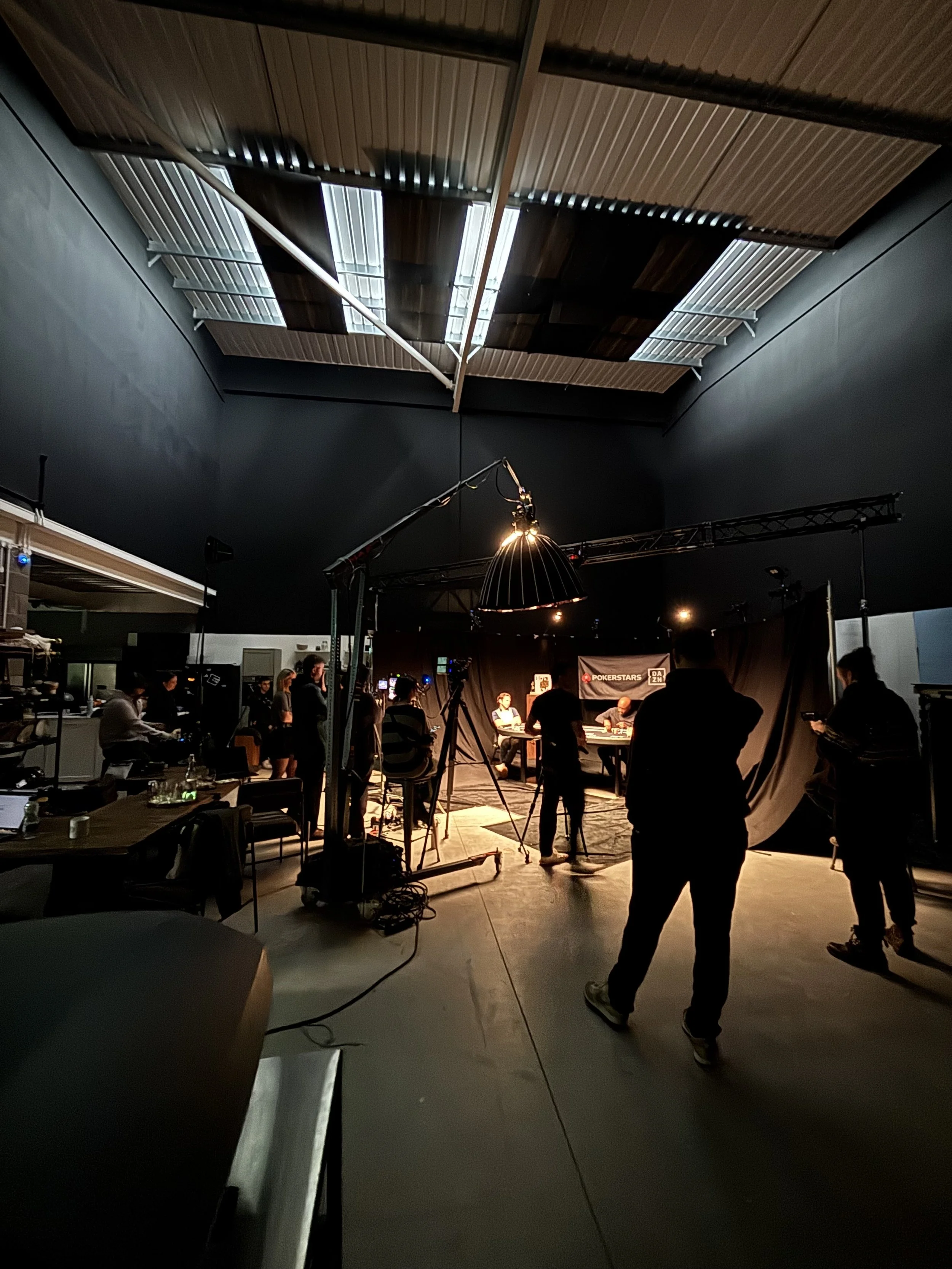 A poker tournament in progress with players seated at tables, cameras, and crew setting up lighting, with a black backdrop displaying poker-related logos, inside a modern venue with a high wooden ceiling.