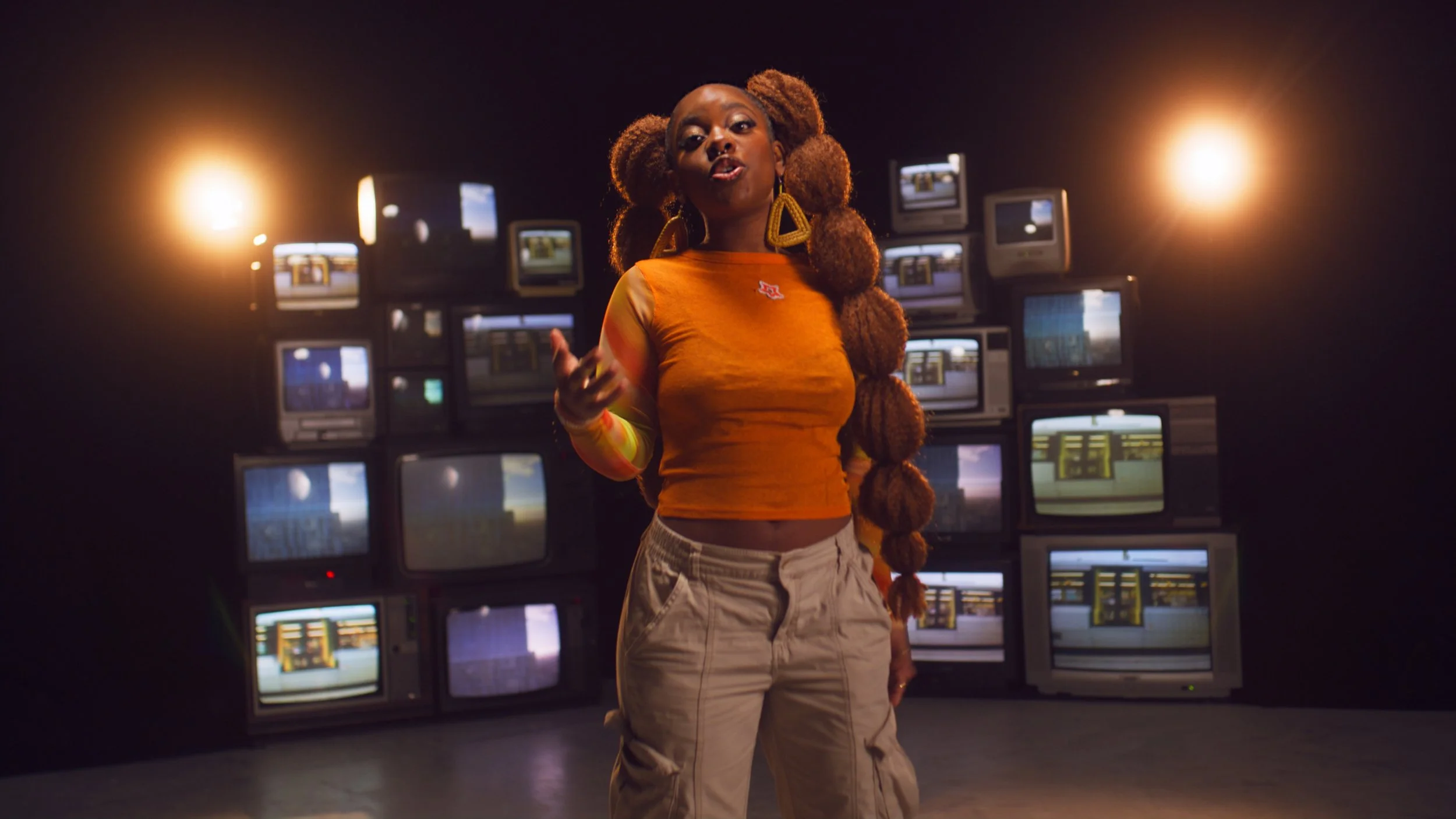 A woman with large curly hair and wearing an orange long sleeve shirt with a small logo, standing in front of a wall of vintage television screens, some displaying various scenes, with bright lights on either side of her.