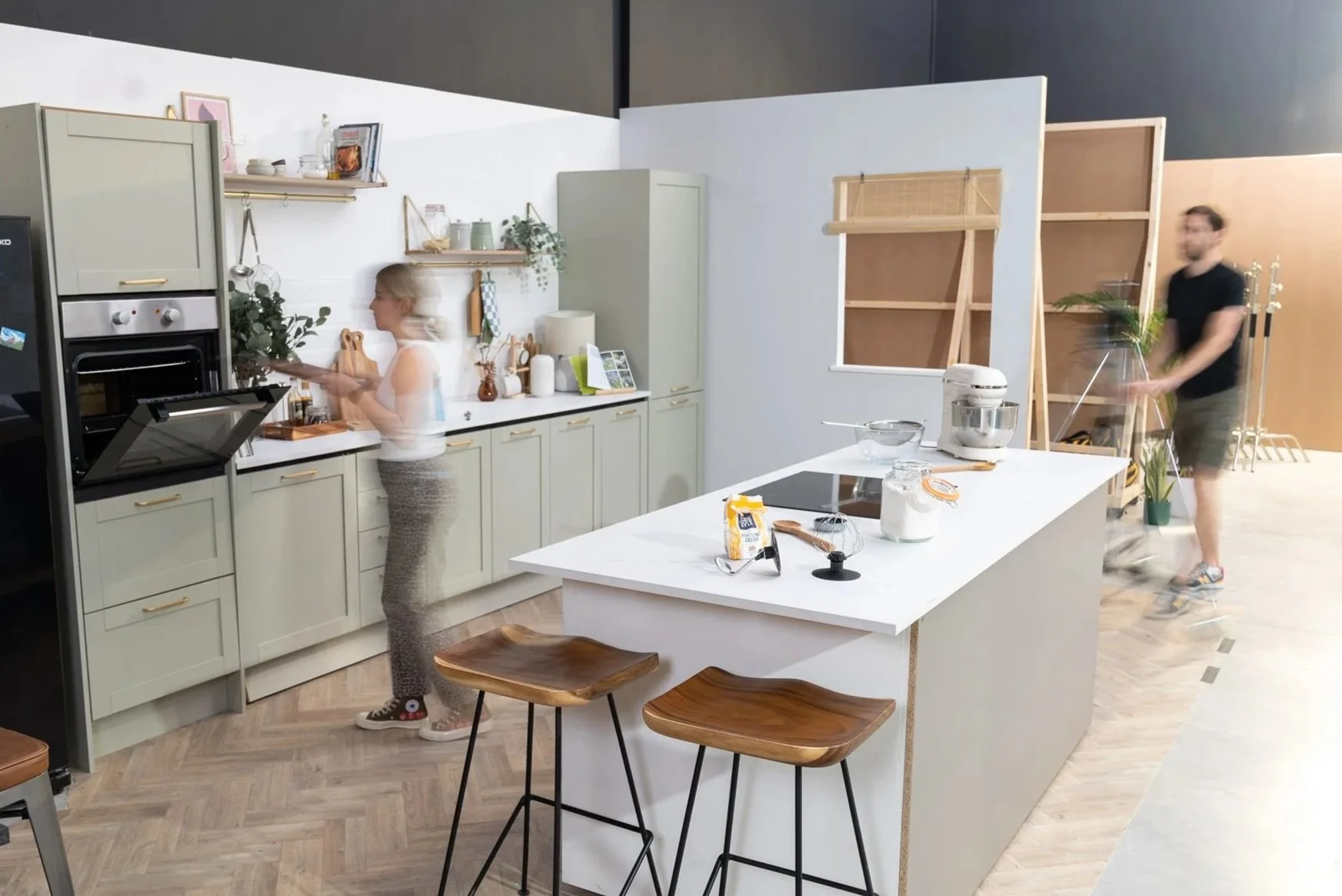 A modern kitchen with a woman cooking at the stove, a man moving a cart in the background, and a white island counter with kitchen utensils and food items on it.