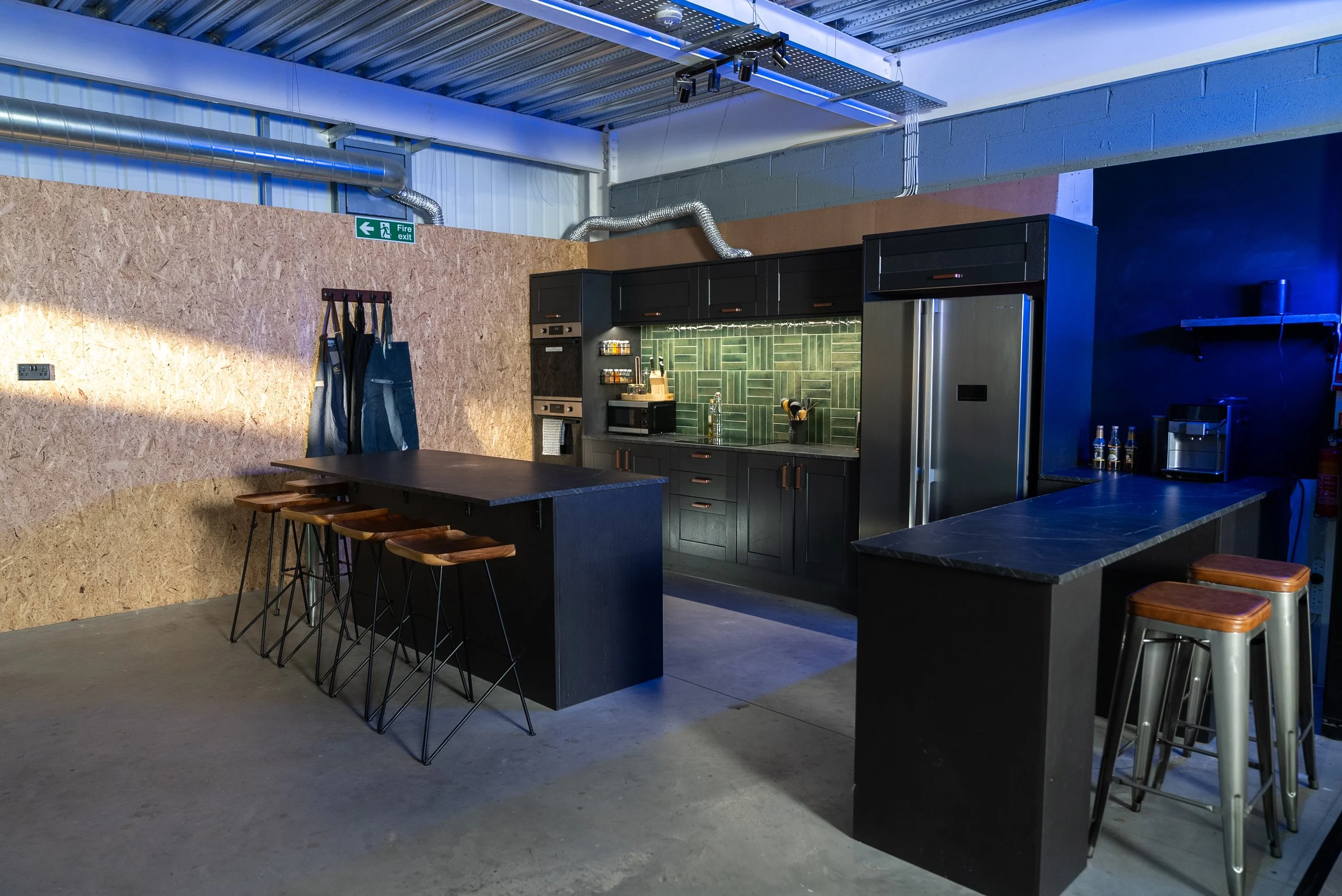 Modern industrial-style kitchen with black cabinets, green tile backsplash, and a mix of seating including bar stools and chairs.