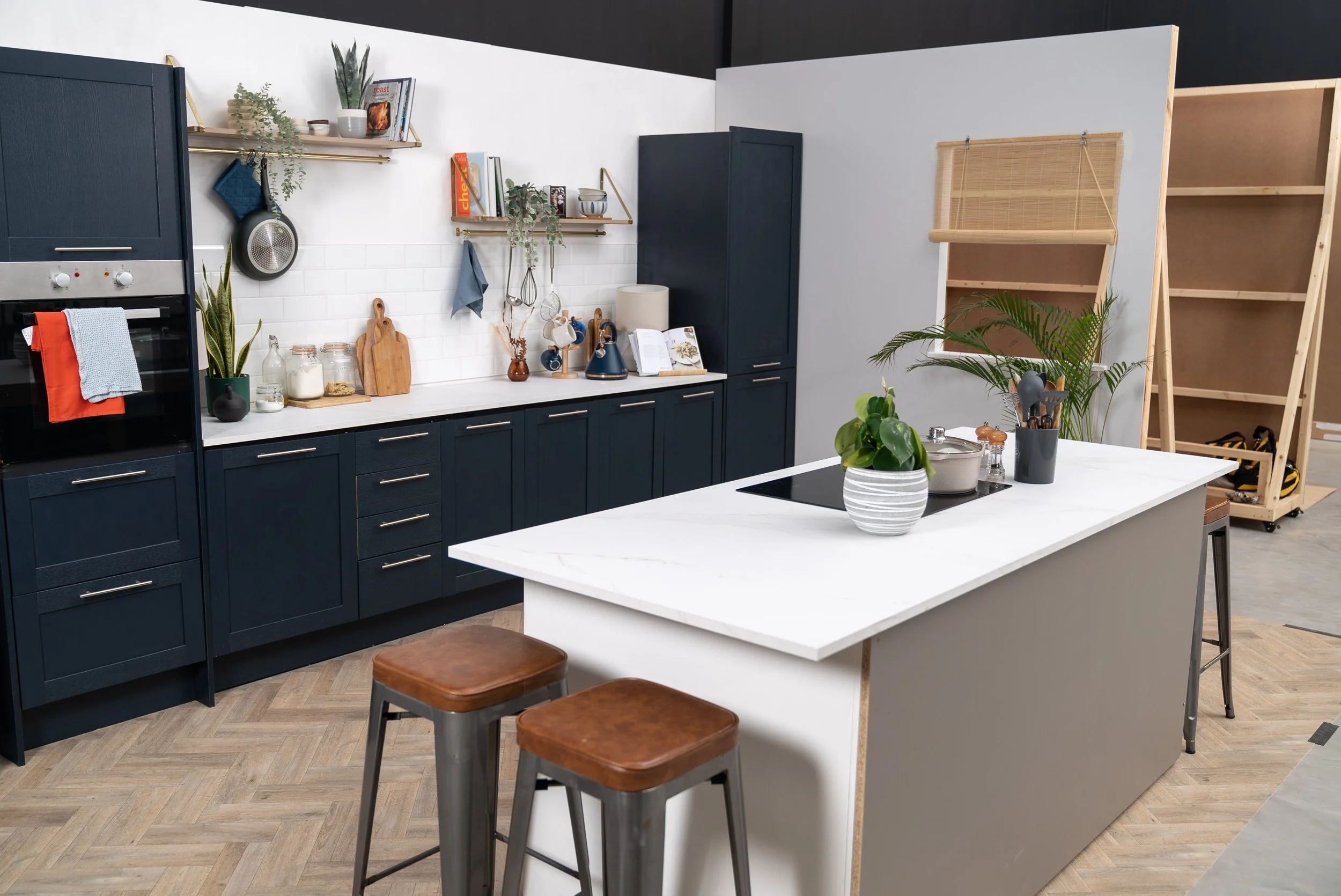 Modern kitchen with navy cabinets, white countertops, open shelves, a white kitchen island with wooden bar stools, and various plants and kitchen tools.