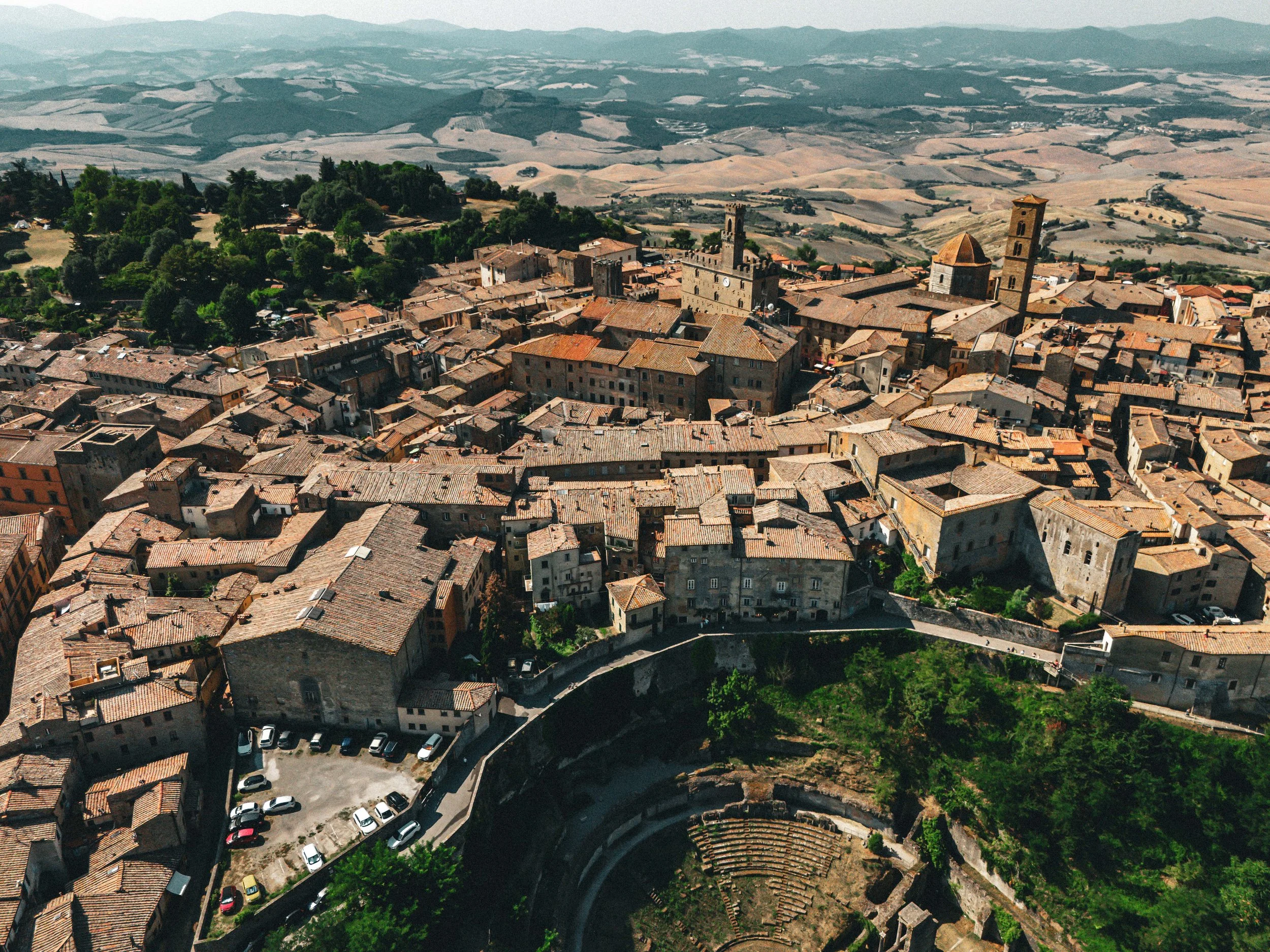 Vue aérienne d'une vieille ville avec des bâtiments en pierre, des églises avec des tours, et un amphithéâtre antique en contrebas, entourée de collines et de champs agricoles.