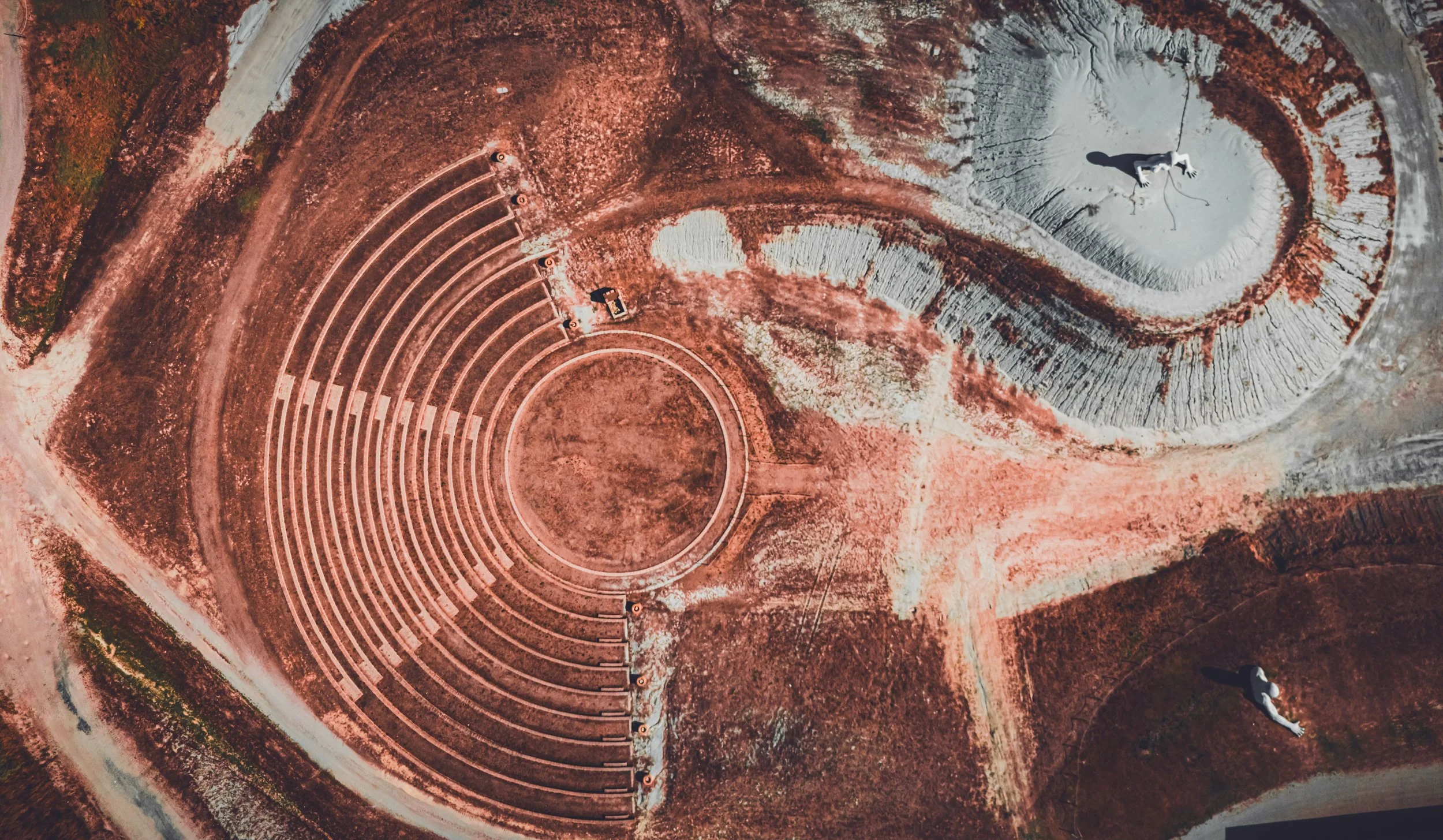 Une image prise d'en haut montrant un amphithéâtre en forme de demi-cercle avec des sièges en pierre rouges, un espace central en gravier, et deux personnes dans des zones différentes du site, l'une à proximité de la partie supérieure et l'autre sur la partie inférieure.