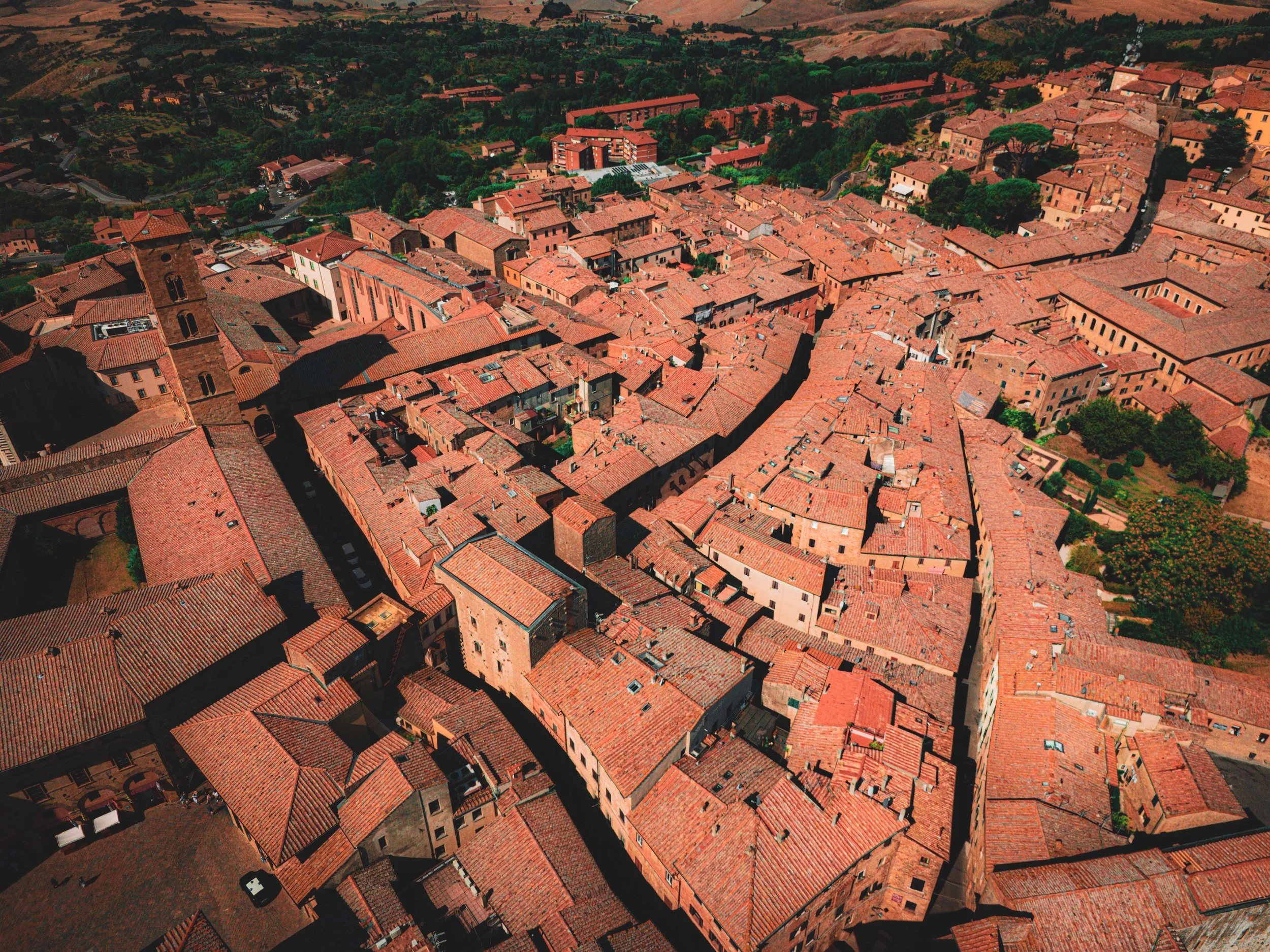 Vue aérienne d'une vieille ville avec des toits en tuiles rouges, des ruelles étroites et des bâtiments anciens, entourée de zones vertes et de collines.