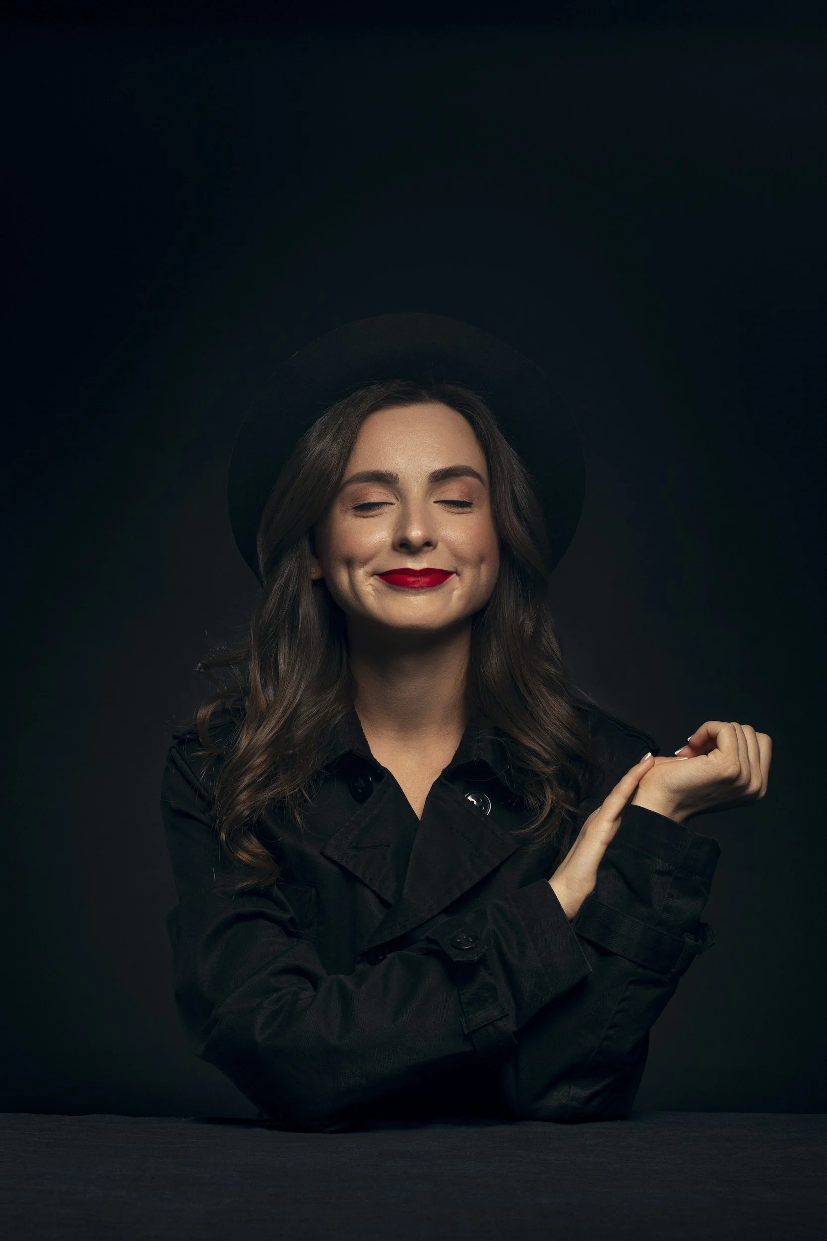 Junge Frau mit dunklem Hut, rotem Lippenstift und schwarzen Jacke, die lächelnd mit verschränkten Armen vor dunklem Hintergrund sitzt.