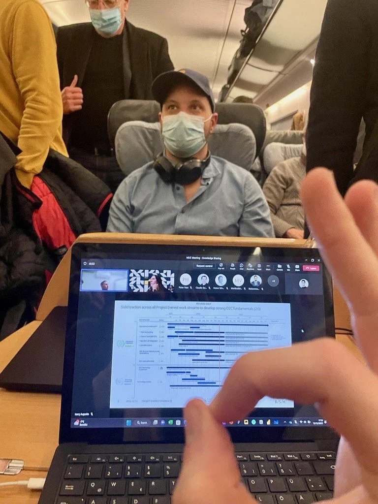Ein Mann sitzt in einem Flugzeug, trägt eine Maske, Kopfhörer und eine Mütze, während um ihn herum andere Passagiere stehen. Vor ihm auf dem Tisch ist ein Laptop, der eine Präsentationsfolie zeigt.