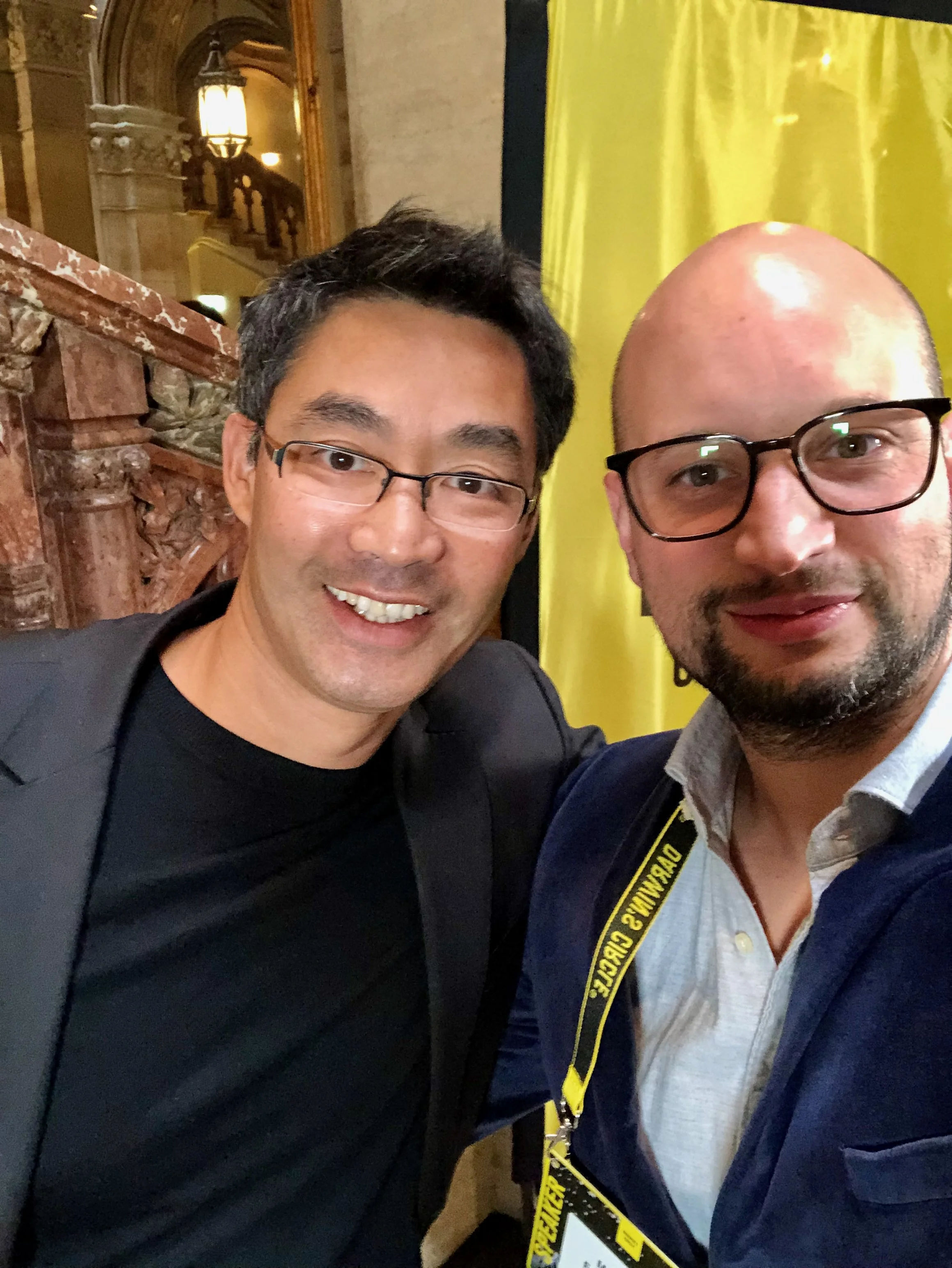 Zwei Männer machen ein Selfie in einem Gebäude mit viktorianischer Architektur, einer trägt eine schwarze Brille, der andere eine dunkelblaue Jacke und eine Kamera um den Hals.