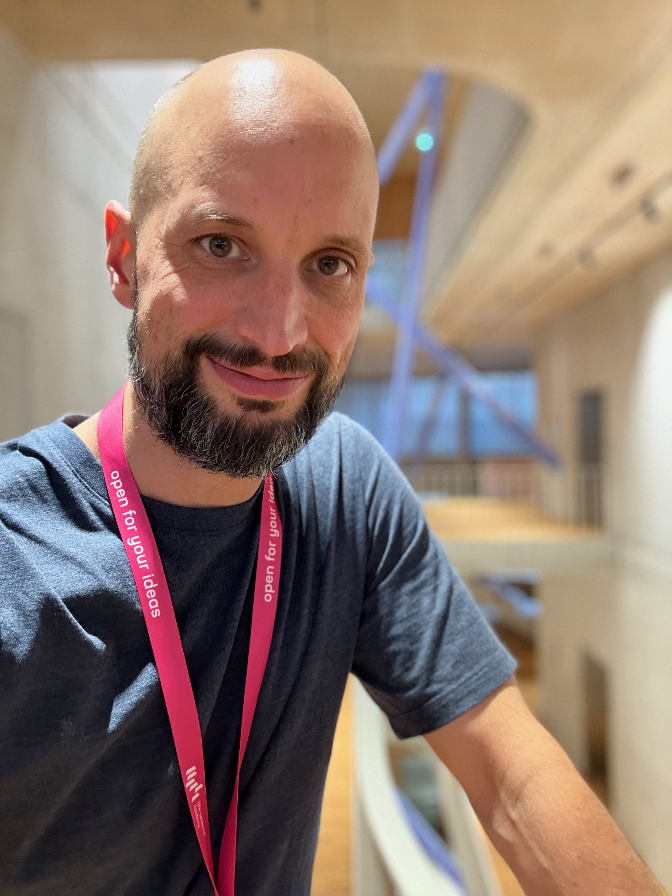 Ein Mann mit Bart und Glatze trägt ein schwarzes T-Shirt und eine pinkfarbene Lanyard. Er steht in einem Holzhaus oder einer Holzkonstruktion mit sichtbaren Holzbalken im Hintergrund. Er lächelt leicht in die Kamera.