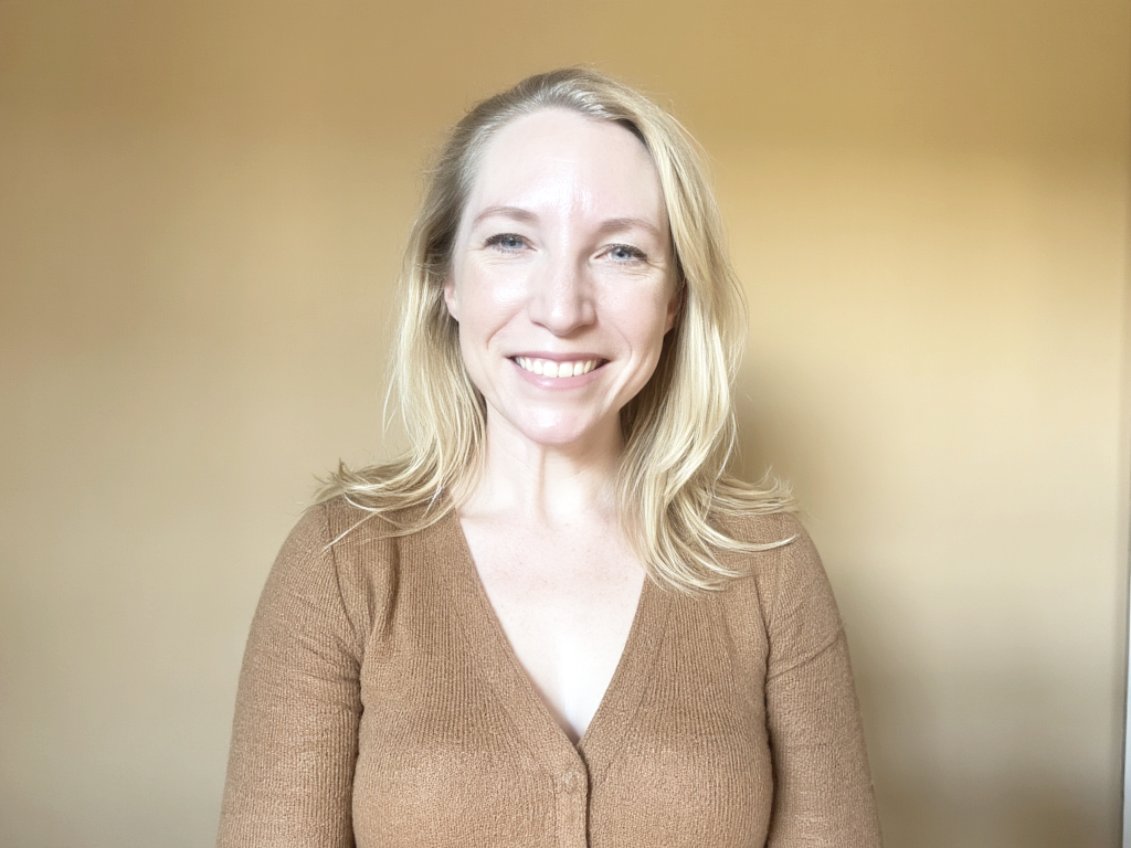 A smiling leader wearing a brown sweater standing against a beige background.