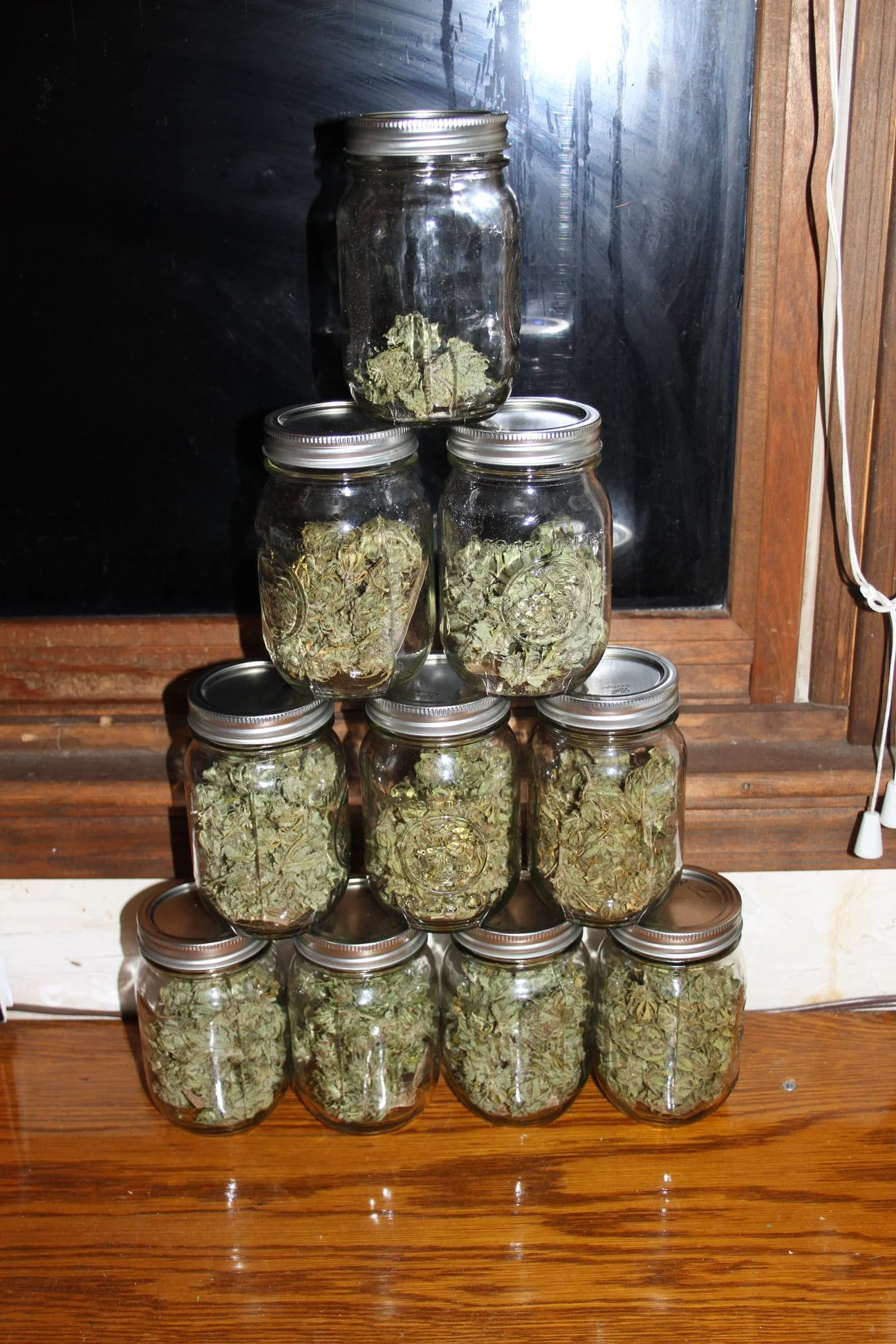 A pyramid of nine glass jars filled with dried cannabis buds, stacked on a wooden surface in front of a window.