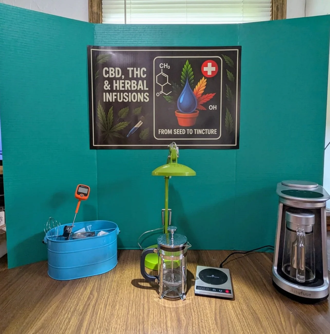 Display booth with equipment for CBD, THC, and herbal infusions, featuring a sign that says "From Seed to Tincture" with symbols of chemical structures, a dropper, and cannabis leaves, along with a small table, a digital scale, and a vaporizer.