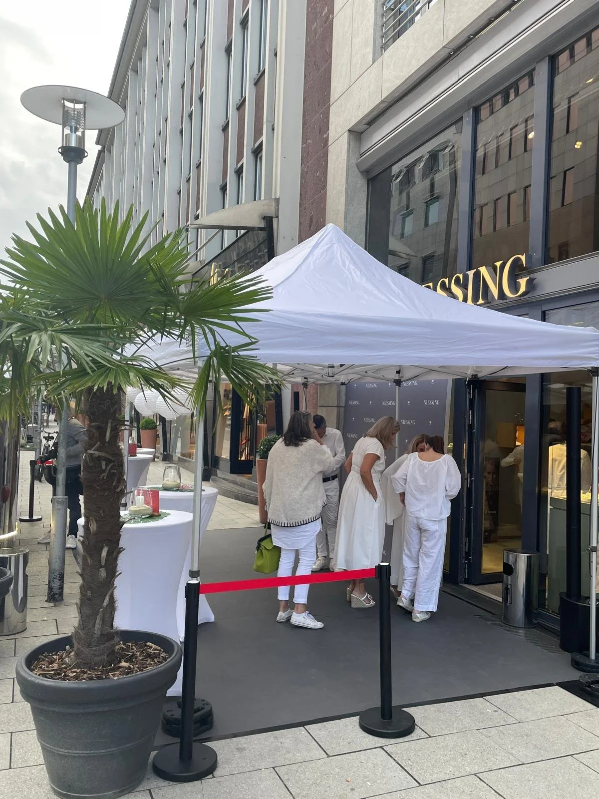 Menschen warten vor einem Geschäft unter einem weißen Zelt auf dem Bürgersteig.