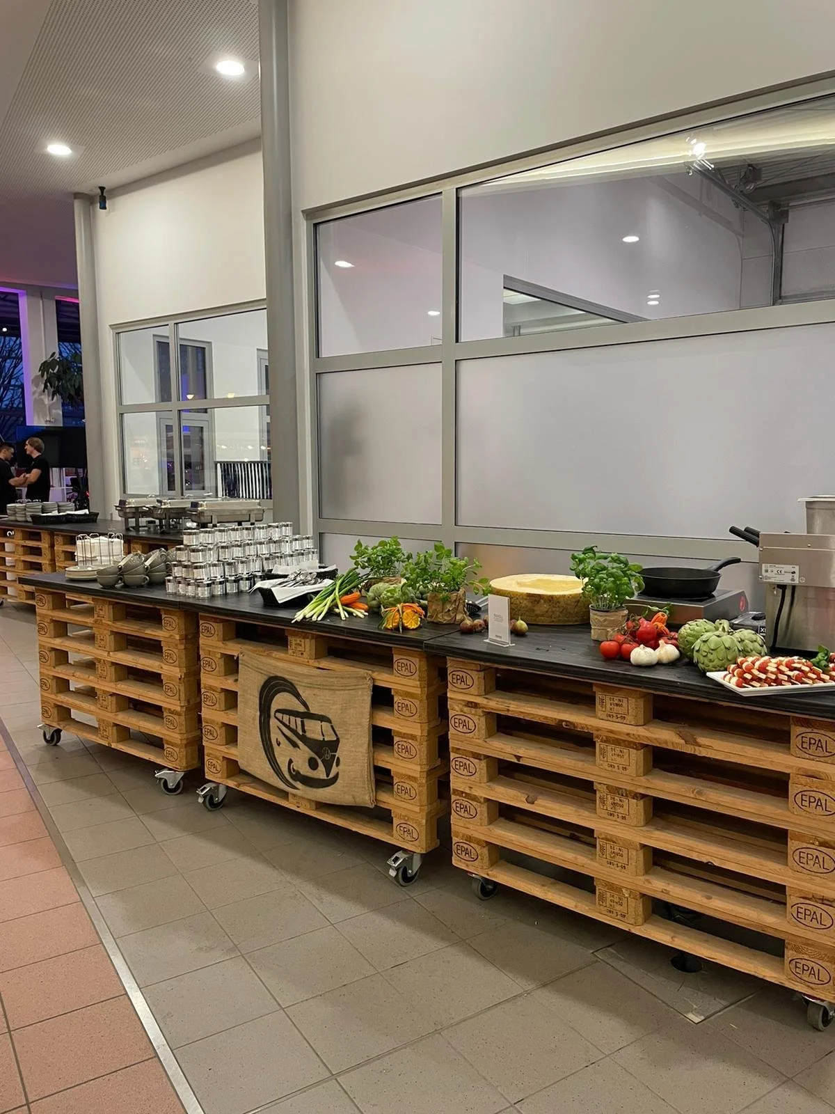 Buffet-Tisch mit Salat, Gemüse und Küchenutensilien in einem modernen Raum, aufgebaut auf Paletten.