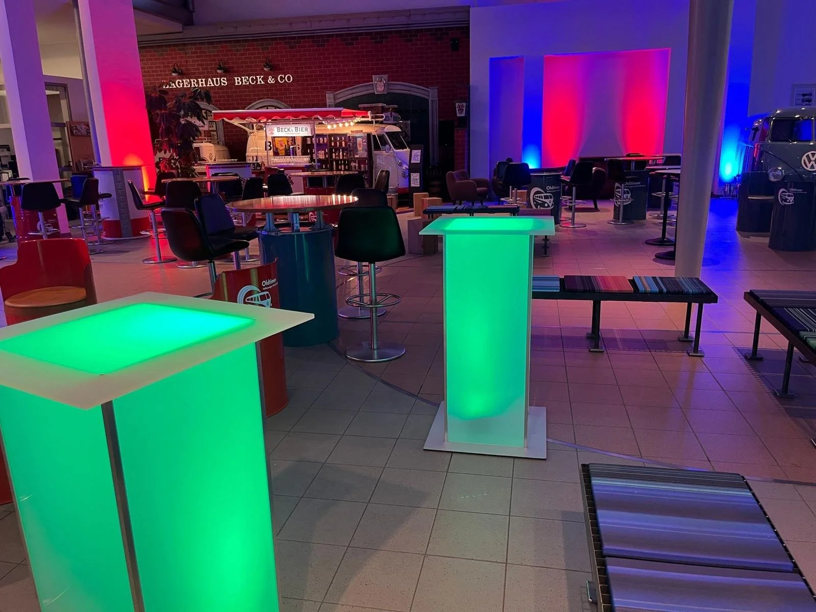 Leeres Veranstaltungsraum mit bunten Akzentbeleuchtung, Stehtischen, Bänken und einem Stand mit Getränkeauto.