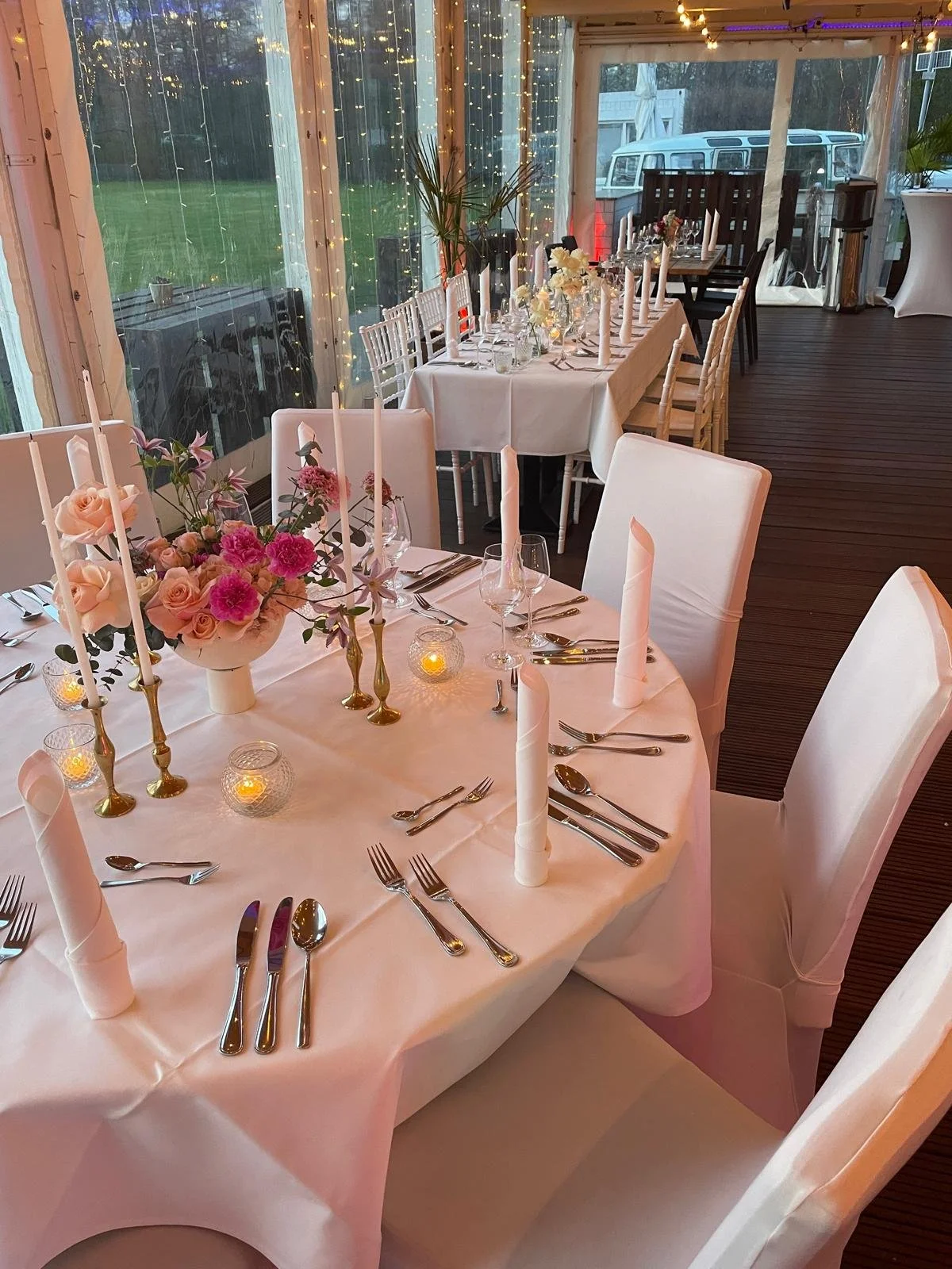 Elegante festliche Tafel mit weißen Tischtüchern, Blumenarrangements, Kerzen, und Besteck in einem festlich geschmückten Raum mit Lichterketten.