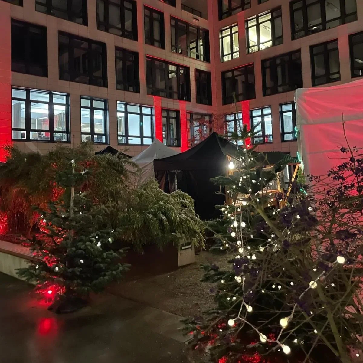 Weihnachtlich geschmückter Innenhof mit Lichterketten, Tannenbäumen und Zelten vor einem modernen Gebäude mit großen Fenstern bei Nacht.