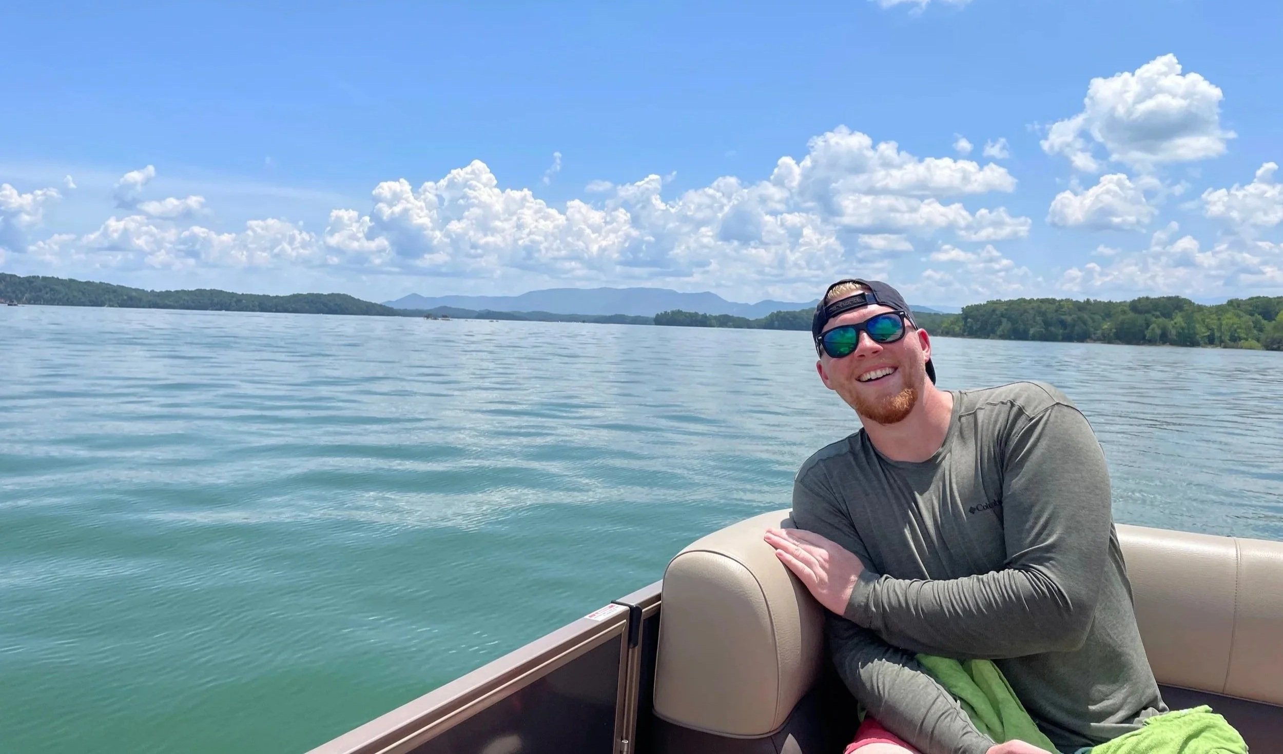 A man smiling on a boat with a lake and mountains in the background under a partly cloudy sky.